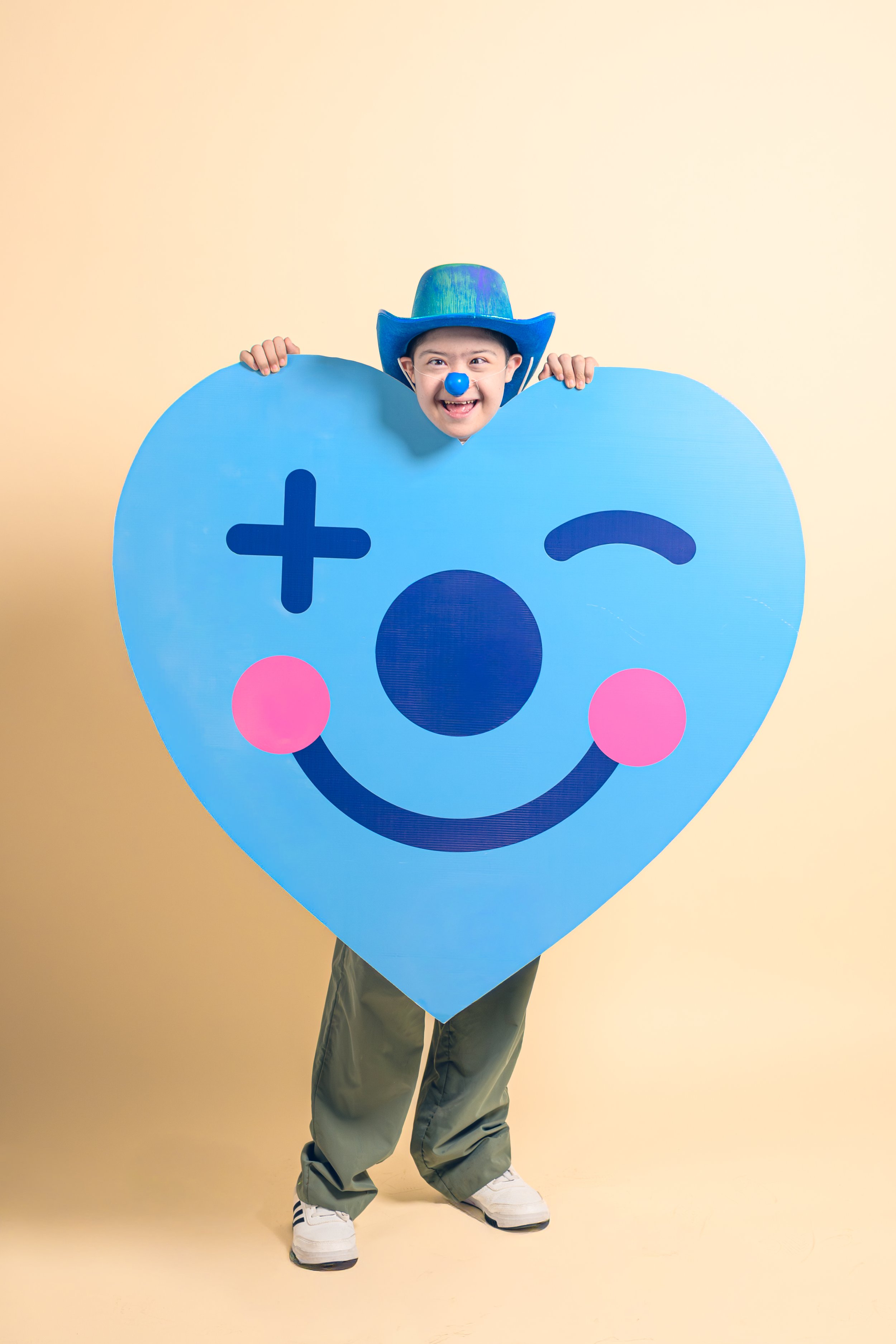 Un niño sonriente con sombrero azul y nariz azul, sosteniendo un cartel en forma de corazón azul con una cara de emoticón sonriente y ojos cerrados, en un fondo color crema.