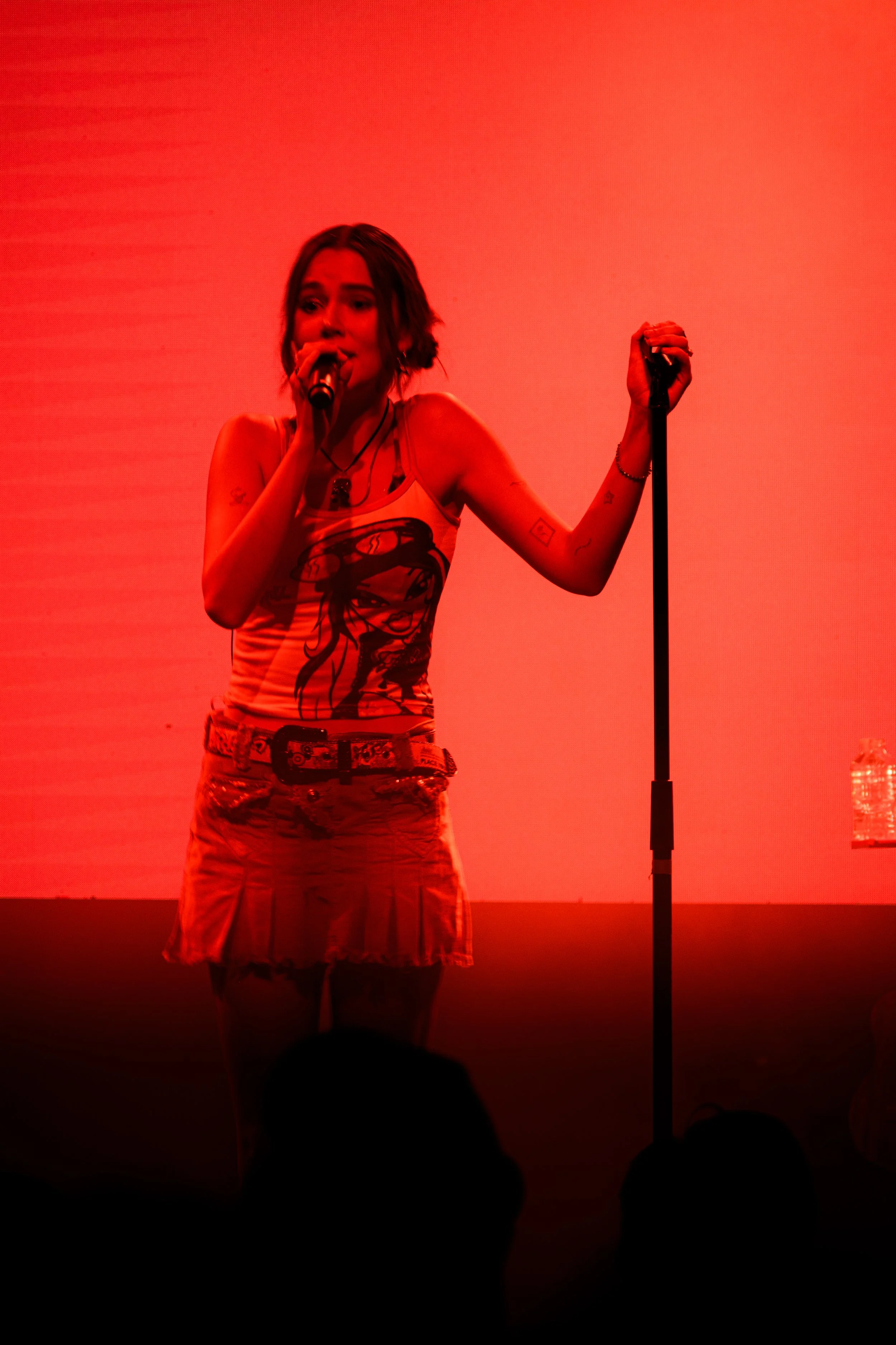 Joven cantante en escenario con iluminación roja, sosteniendo micrófono y posando con una mano en un micrófono de pie, usando camiseta con personaje y falda