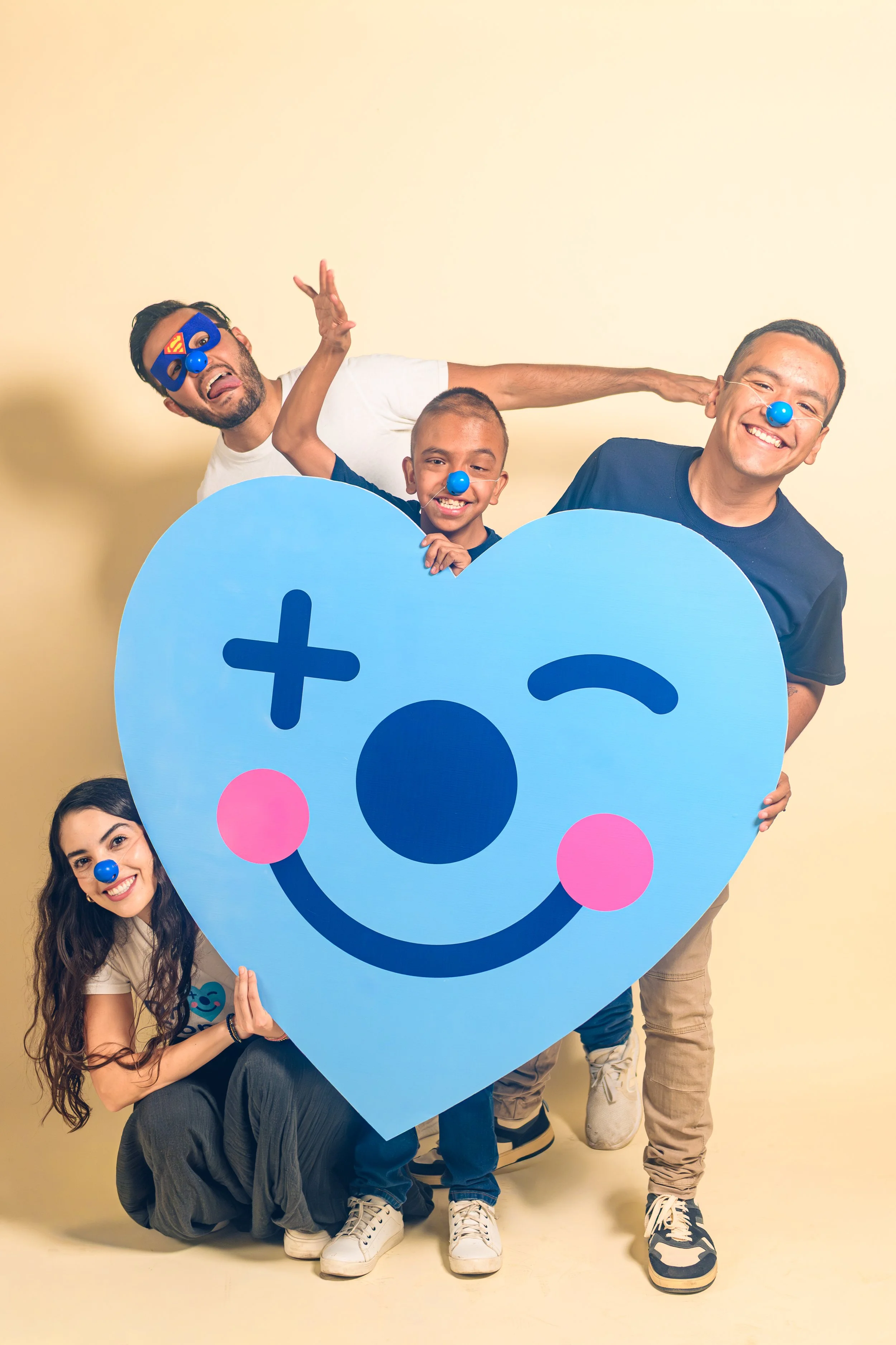 Grupo de cinco personas jóvenes sonriendo y usando narices de payaso, posando con un cartel grande en forma de corazón azul con una cara sonriente en ella, en un fondo de color beige.