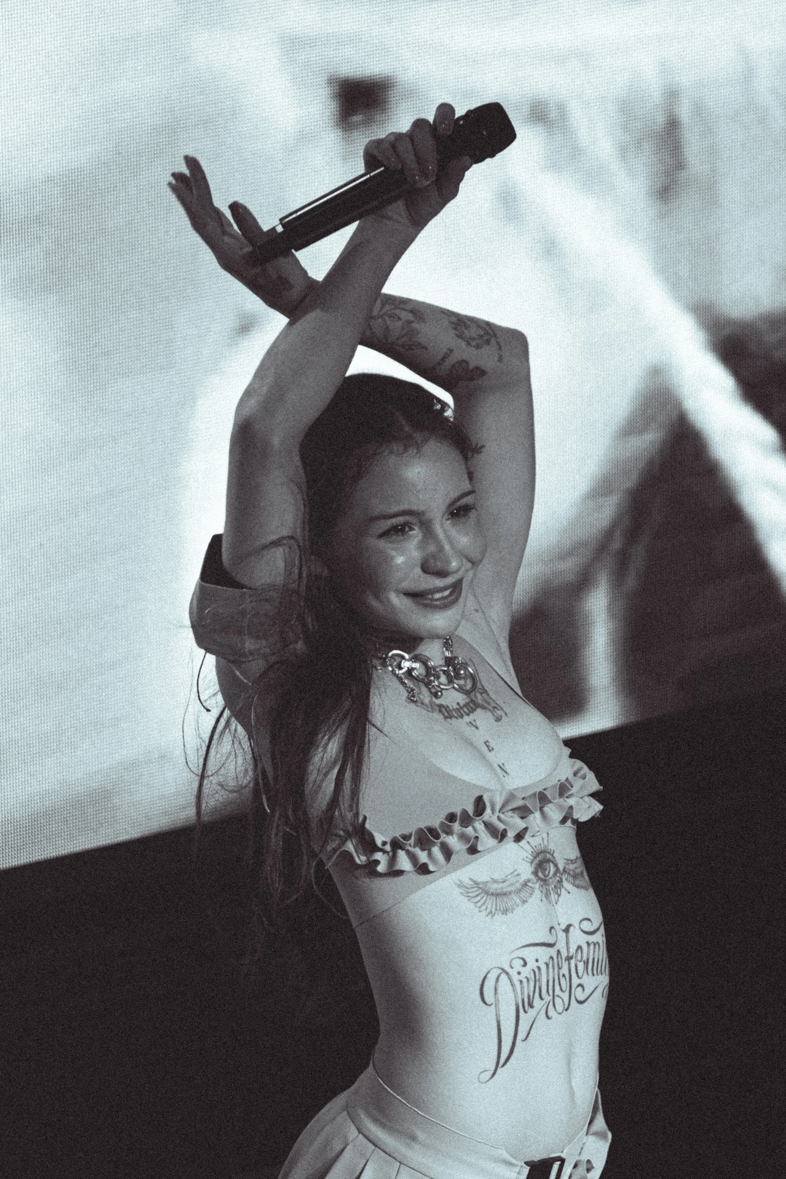 Mujer sosteniendo un micrófono sobre su cabeza en un escenario, sonriendo y luciendo tatuajes y ropa llamativa en un concierto.