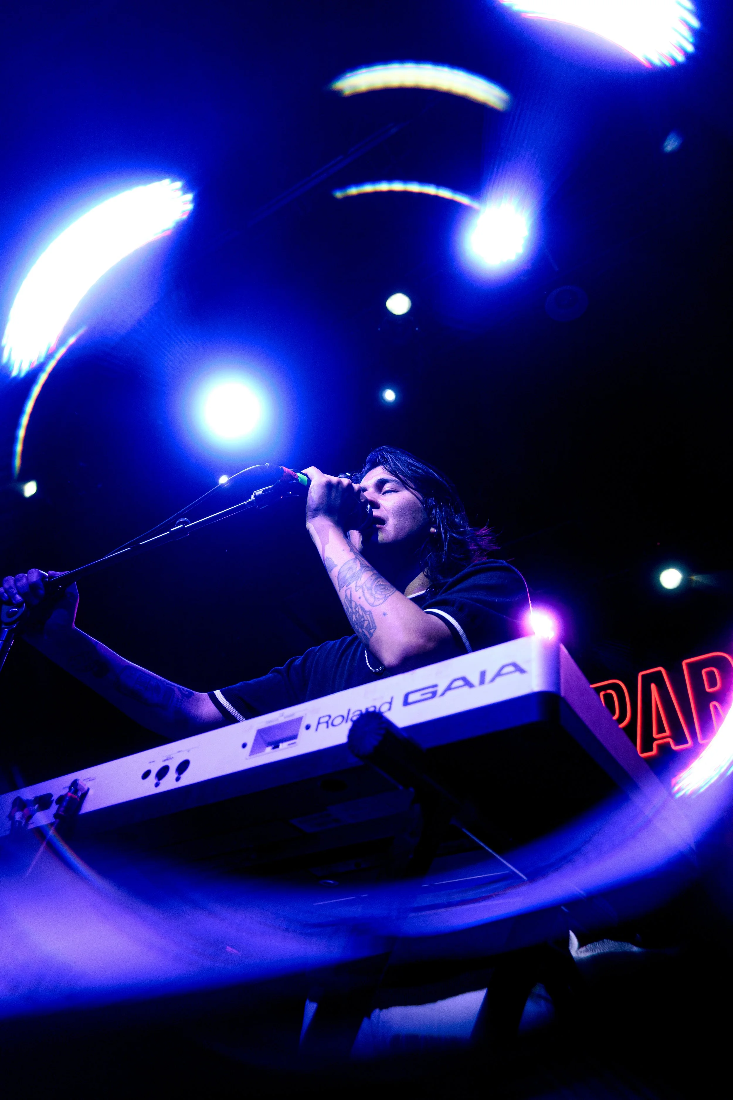 Mujer tocando sintetizador Roland Gaia en escenario con luces azules y neón en el fondo.