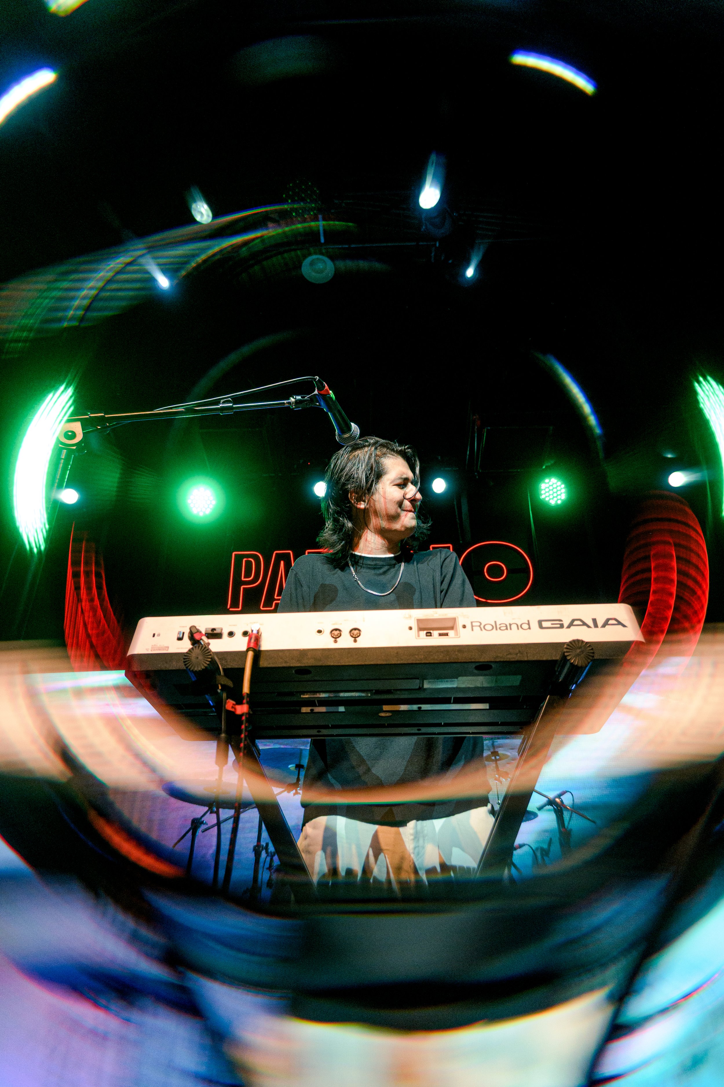 Joven mujer tocando un teclado en un escenario con luces verdes y rojas, vista a través de un cristal o lente distorsionado.