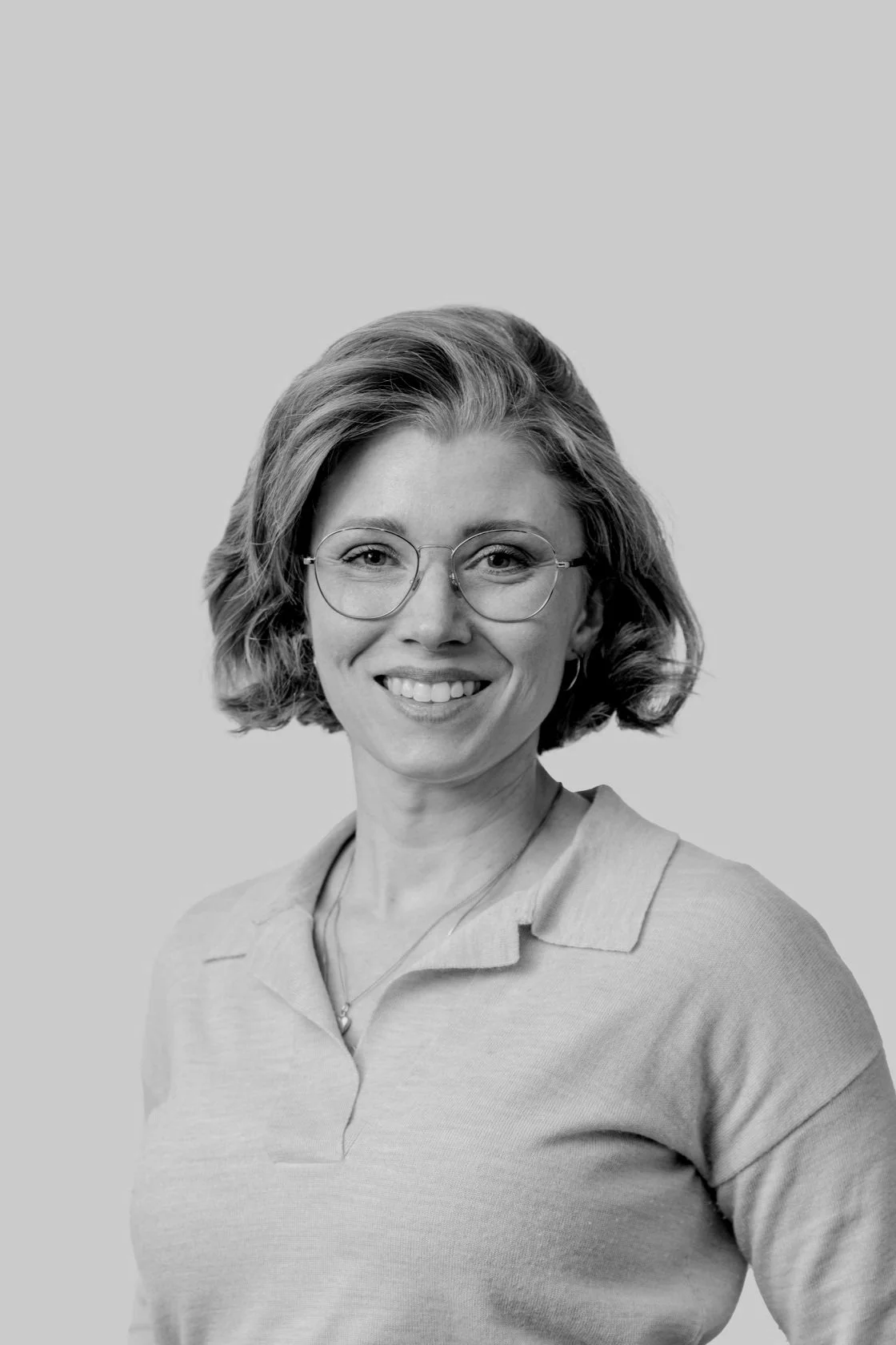 A smiling woman with short wavy hair and glasses, wearing a light-colored polo shirt, posing against a plain light background.