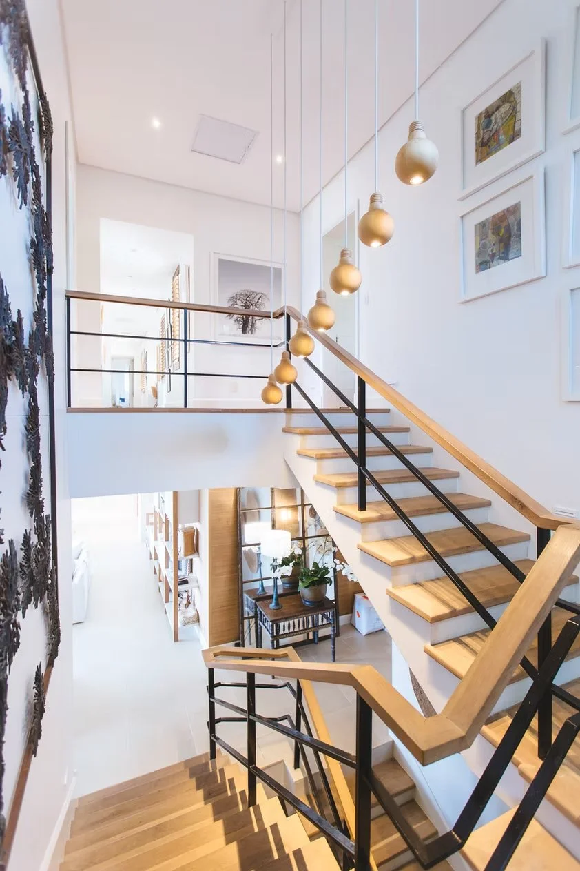 Escaleras de madera en un interior moderno con iluminación colgante en forma de bombillas doradas, paredes blancas con cuadros y una decoración con plantas y una lámpara.