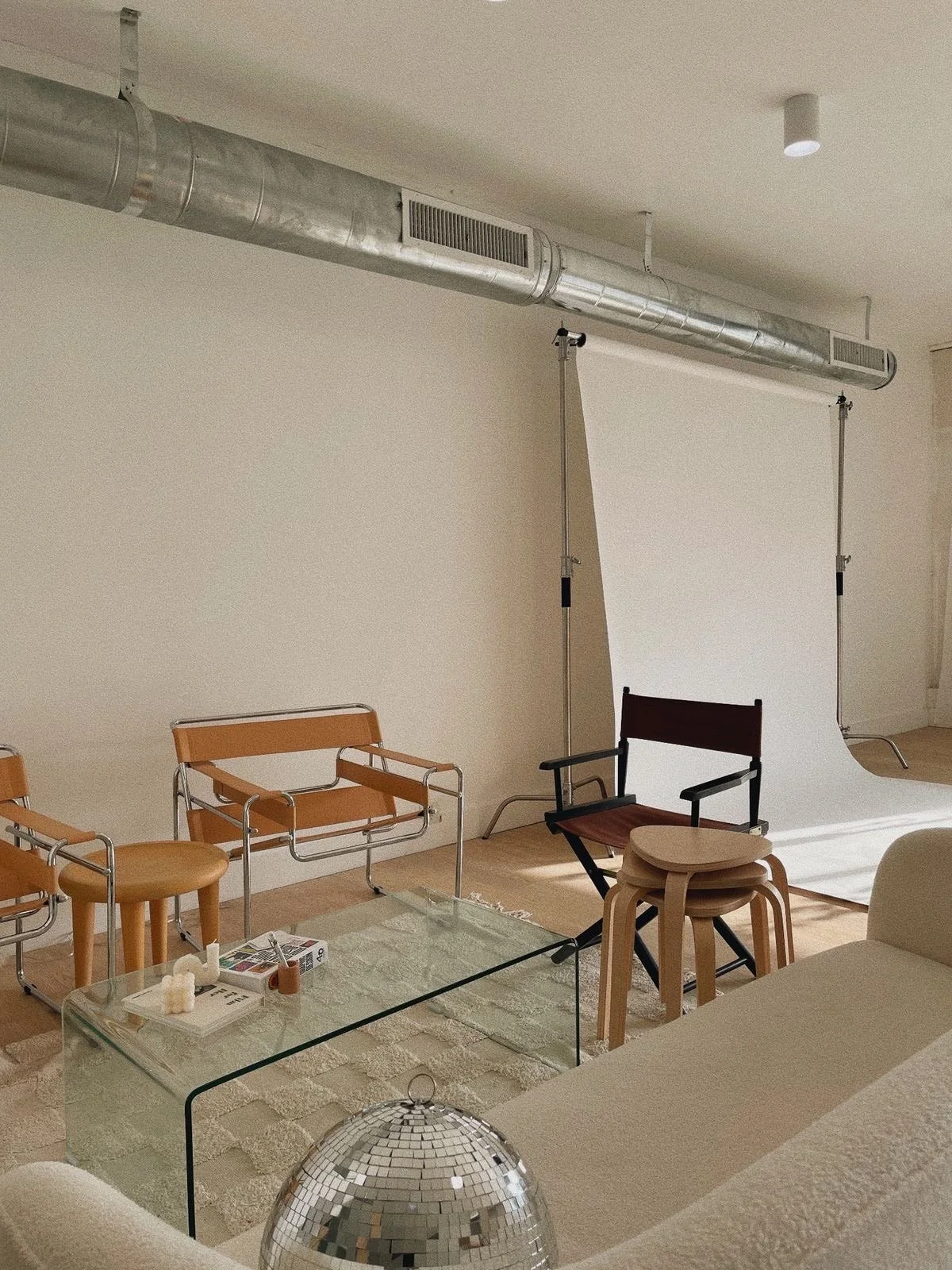 Interior of a room with a white backdrop, chairs, a glass coffee table, and decorative items, with a large silver ventilation duct running along the ceiling.