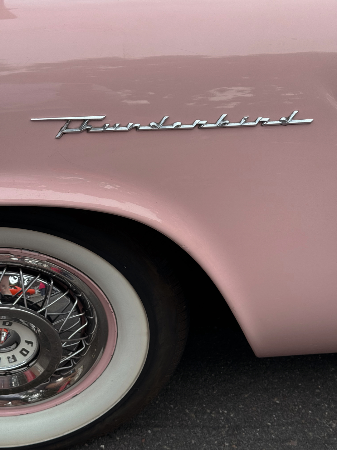 Close-up of a vintage pink car with a chrome 'Thunderbird' emblem on the fender, whitewall tire, and wire-spoke wheel.