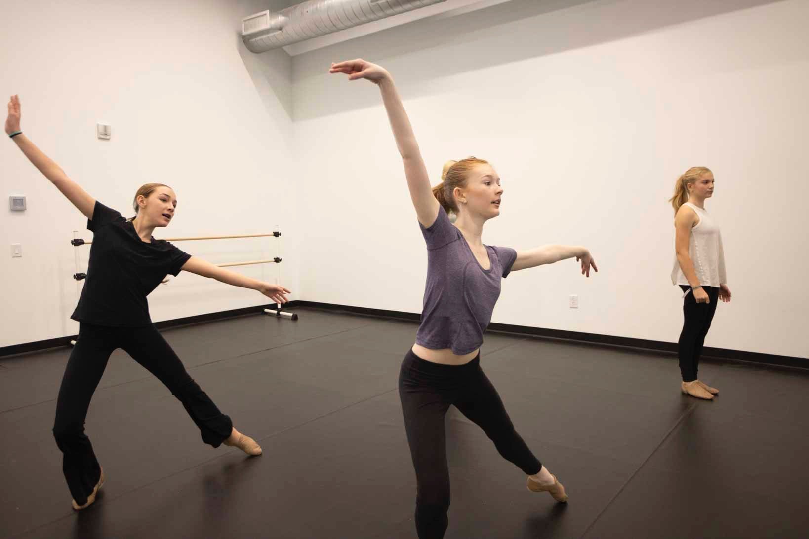Three young women practicing ballet in a dance studio with a black floor and white walls.