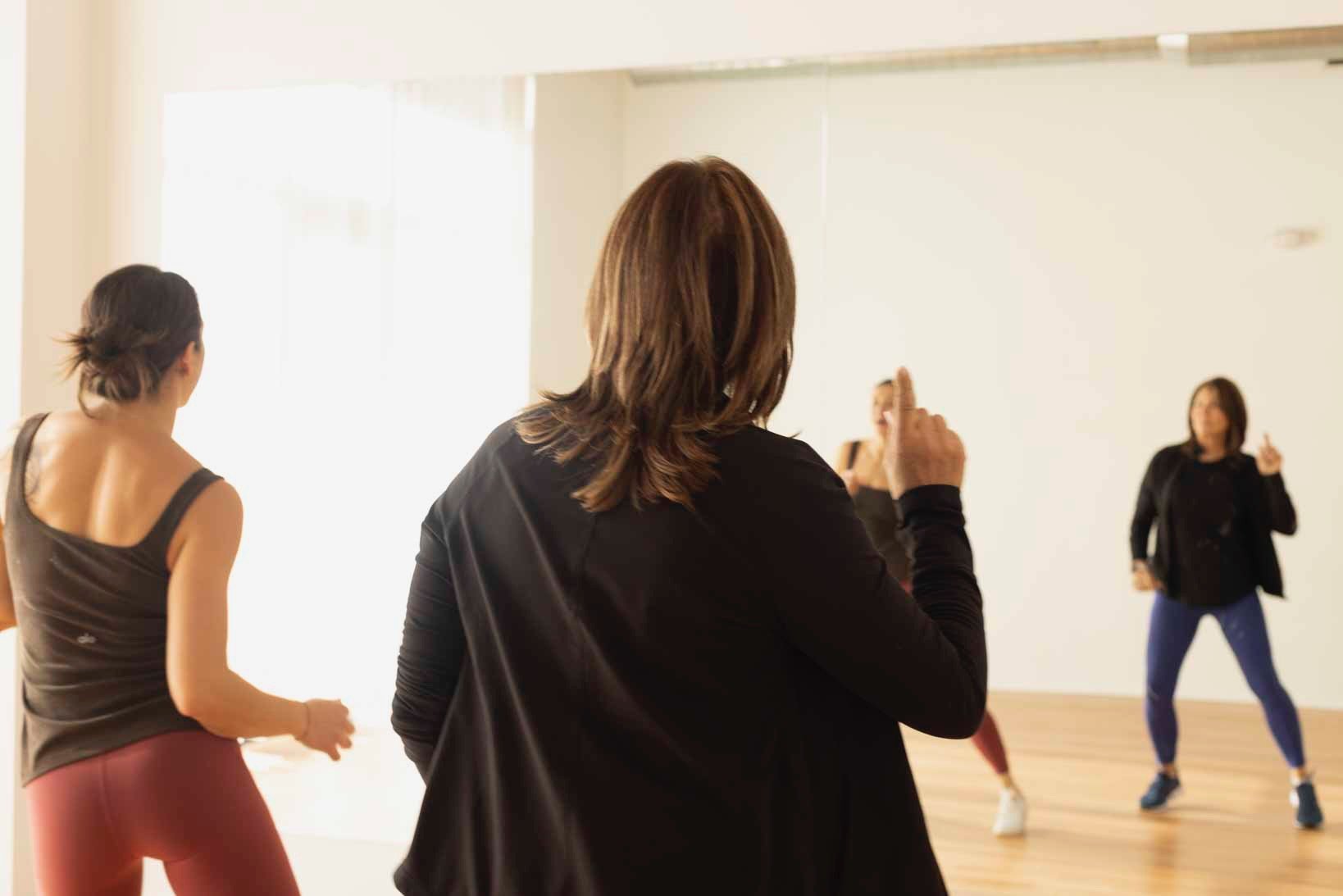 A dance class with four women practicing dance moves in a bright studio with large mirror. One woman in the foreground, with shoulder-length brown hair, appears to be giving instructions or coaching the others.
