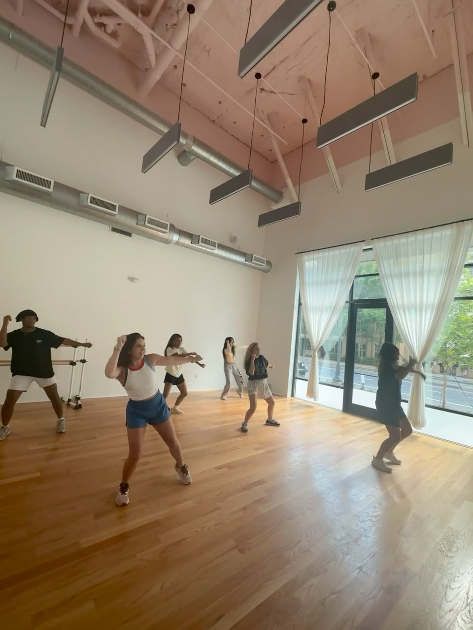 Group of people participating in a dance or exercise class in a spacious room with hardwood floors, large windows with curtains, and ceiling-mounted metal and ductwork.