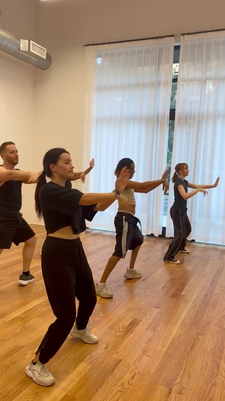 A group of four people practicing a dance or fitness routine in a studio with wooden floors and large windows with white curtains.
