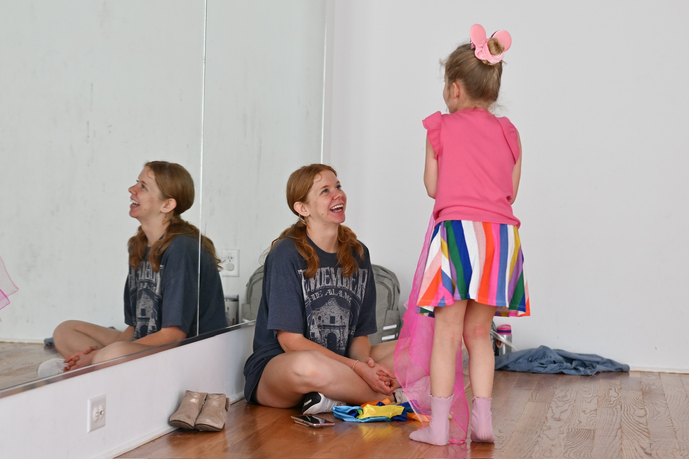 Two women sitting on the floor in front of a mirror, smiling at a young girl standing with her back to the camera, in a bright room.