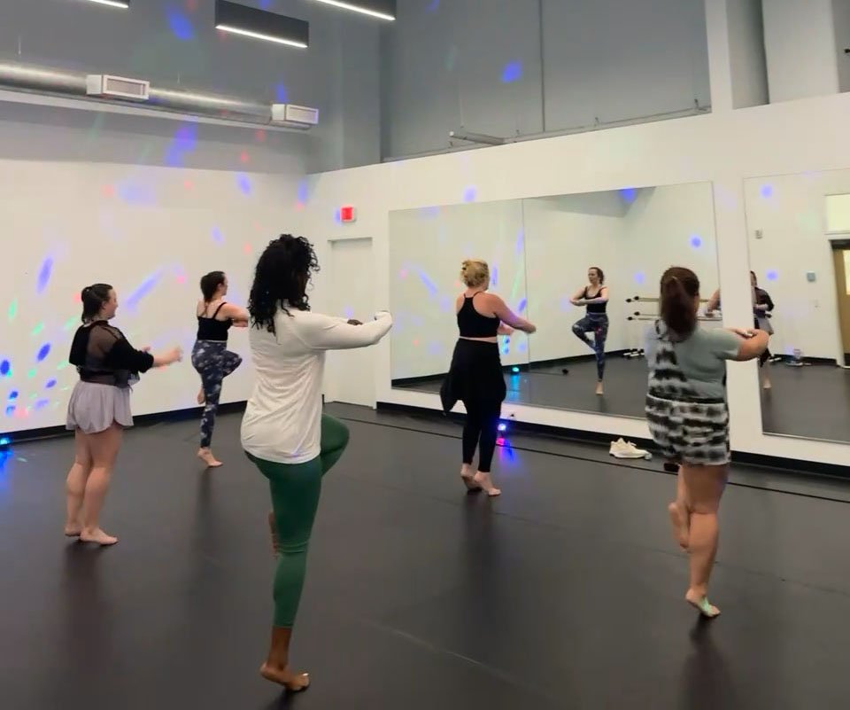 A group of women participating in a dance or fitness class in a studio with large mirrors on the wall and colorful disco lights.