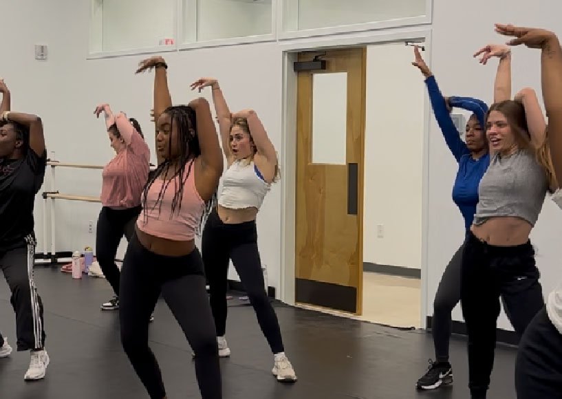 A group of women participating in a dance or fitness class in a studio, raising their arms in the air as part of a choreographed routine.