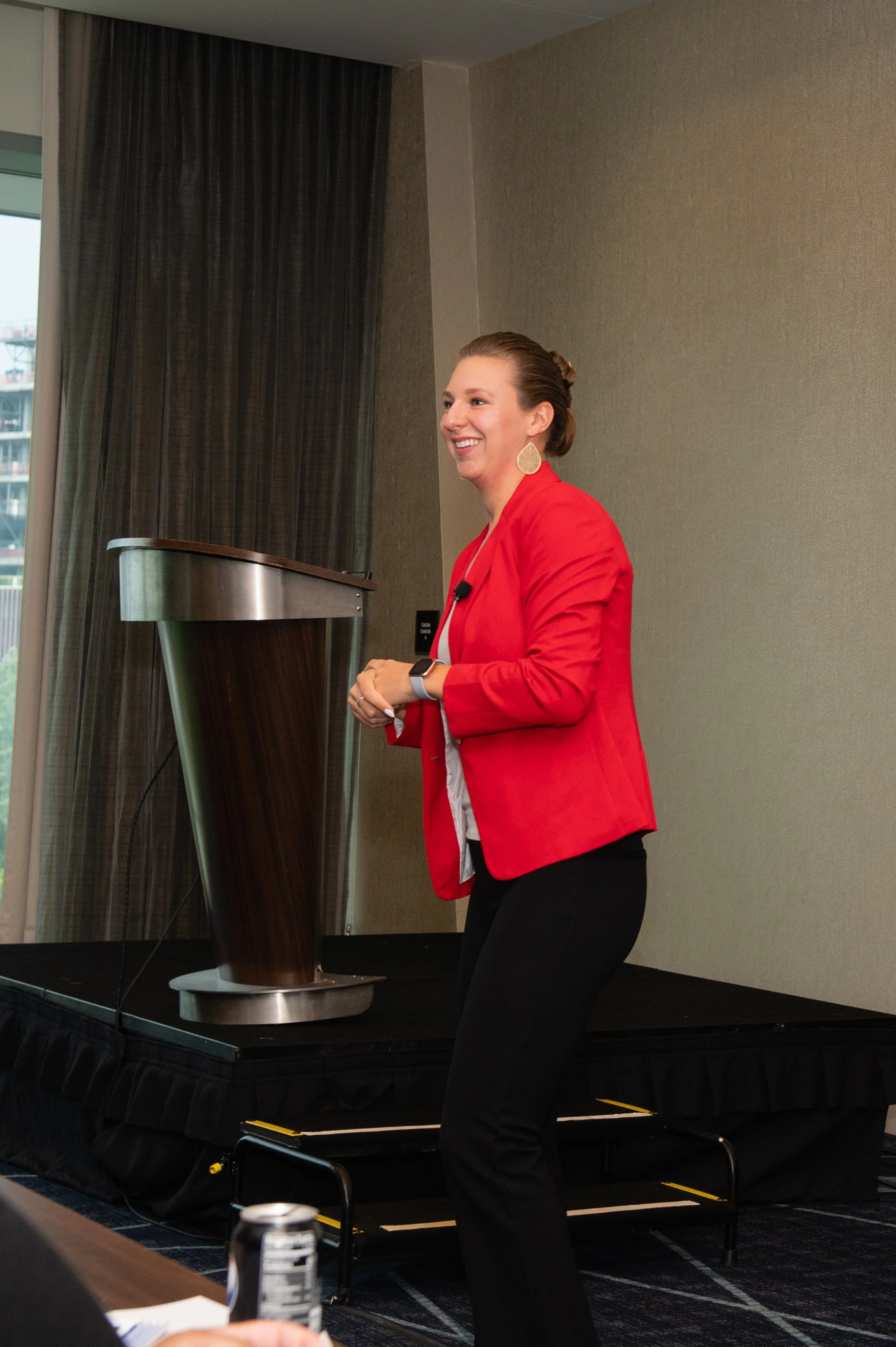 A woman with brown hair tied back, wearing large earrings and a red blazer, is standing on a stage near a podium. She is smiling and appears to be speaking or presenting in a conference room with beige walls, a bookcase window, and a black curtain in the background.