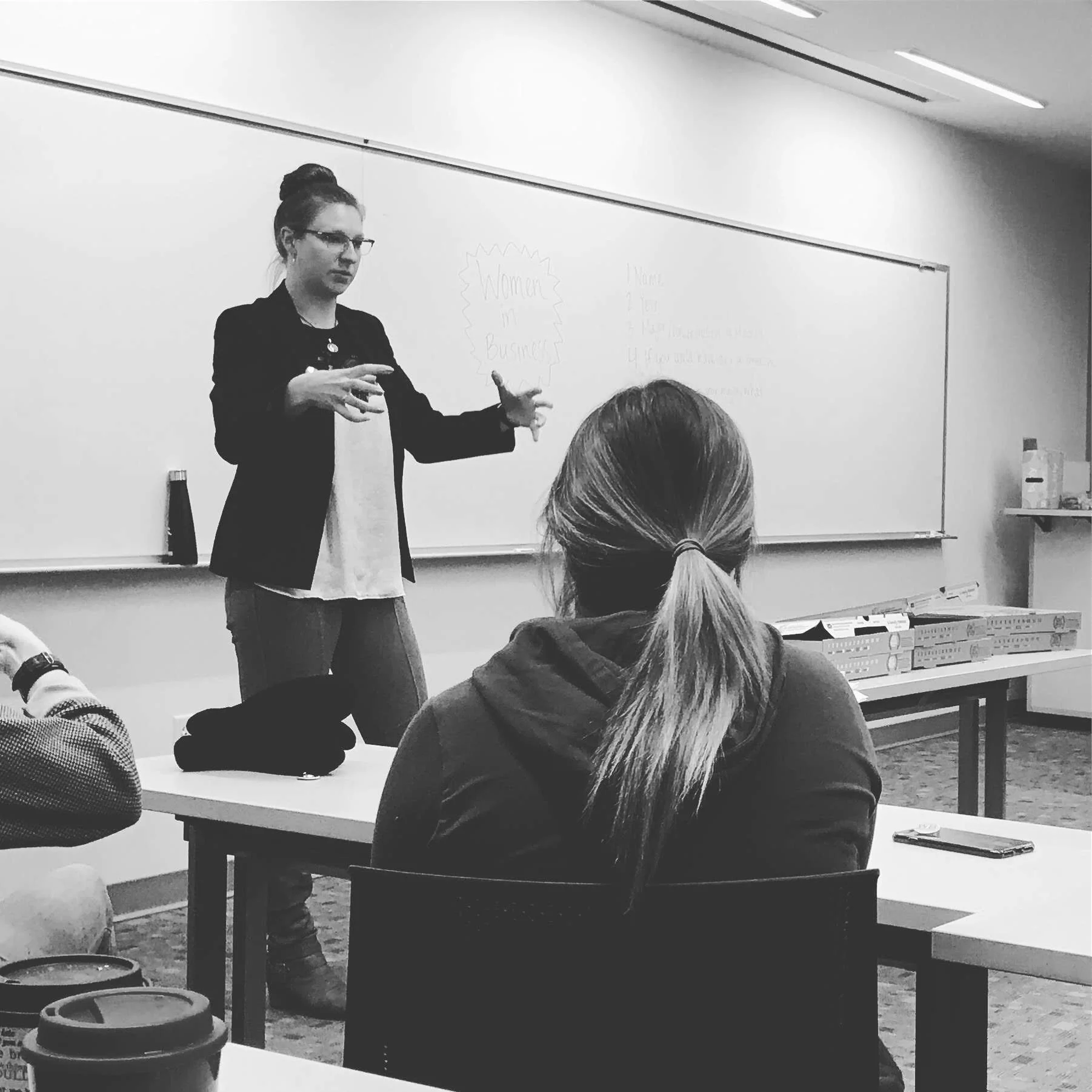 A woman with glasses stands in front of a whiteboard, giving a presentation to a classroom of students seated at desks. The whiteboard has writing and a diagram, and the classroom has bright lighting.