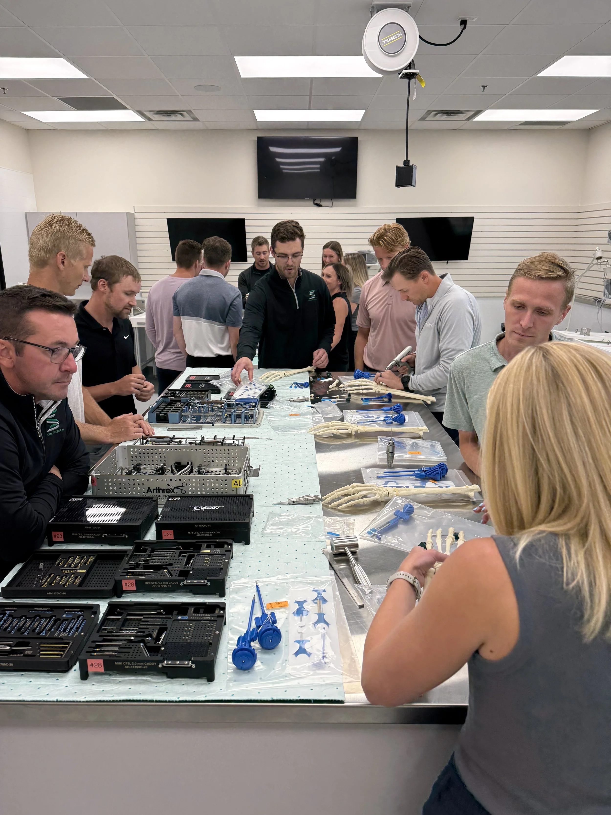 Group of people in a lab, working with medical models of bones, medical instruments, and equipment on a table, in a room with multiple monitors and a ceiling-mounted camera.
