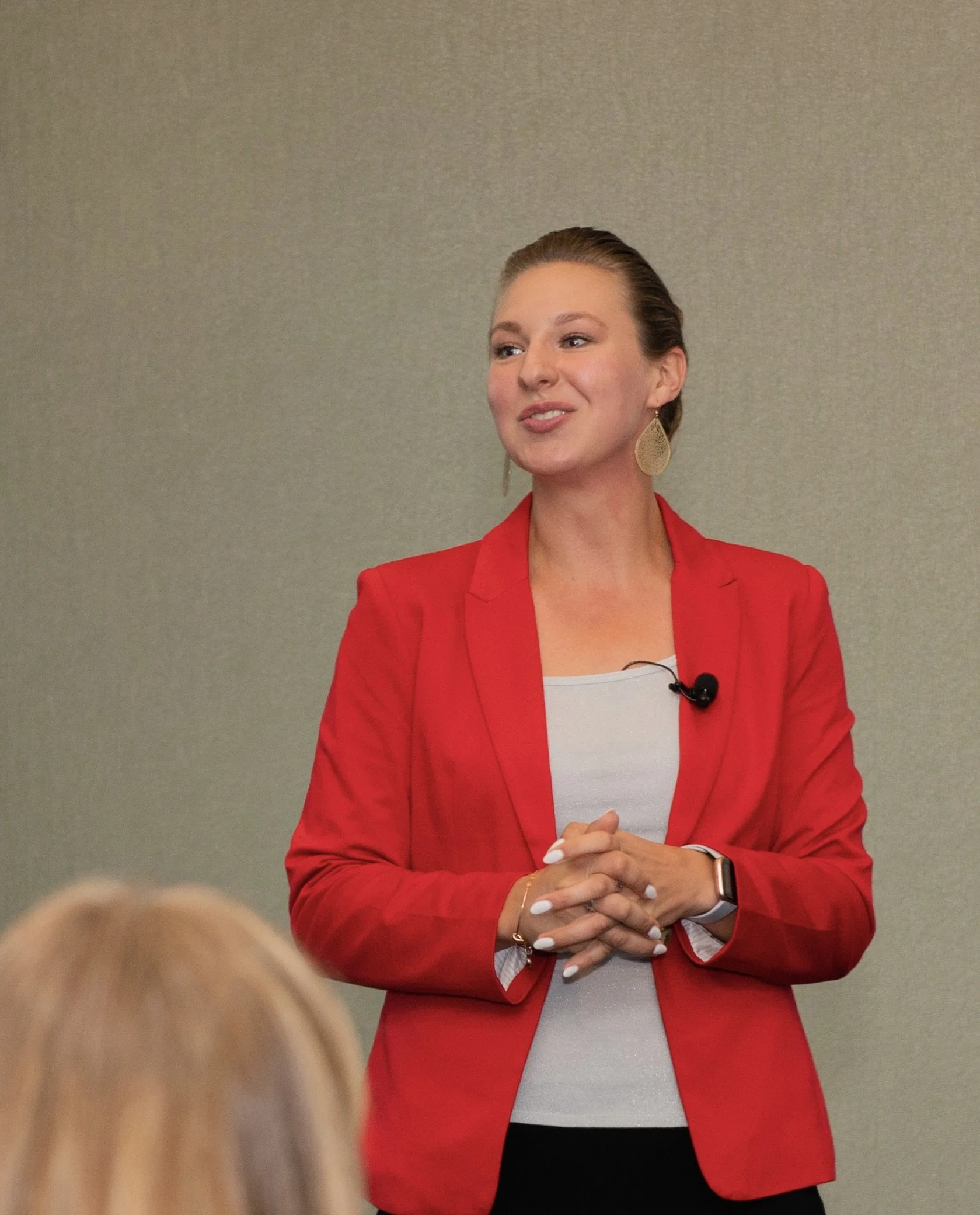 A woman wearing a red blazer and earrings, smiling and speaking at an event, with an audience member in the foreground.