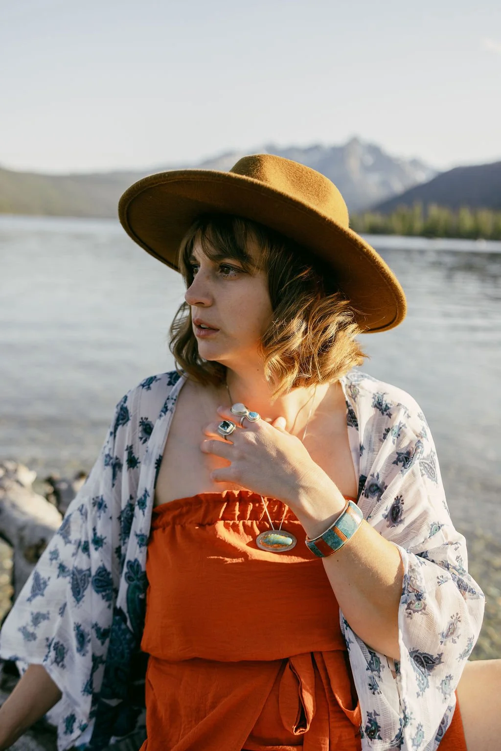 A woman wearing a wide-brimmed brown hat, orange dress, and floral kimono looks thoughtfully into the distance along a river with mountains in the background.