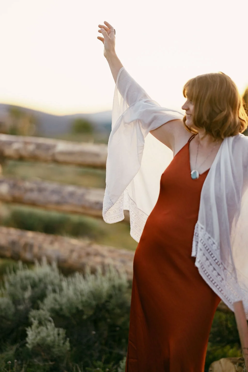 A woman with red hair wearing an orange dress and a white shawl stands outdoors at sunset, with her right arm raised and her left arm by her side.