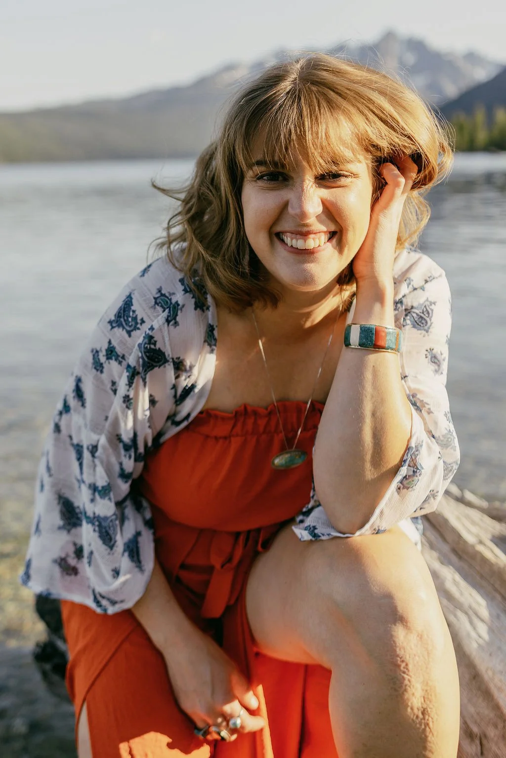 A woman with shoulder-length light brown hair, smiling and leaning on her knee near a body of water with mountains in the background, dressed in a red top, patterned light jacket, and orange pants.