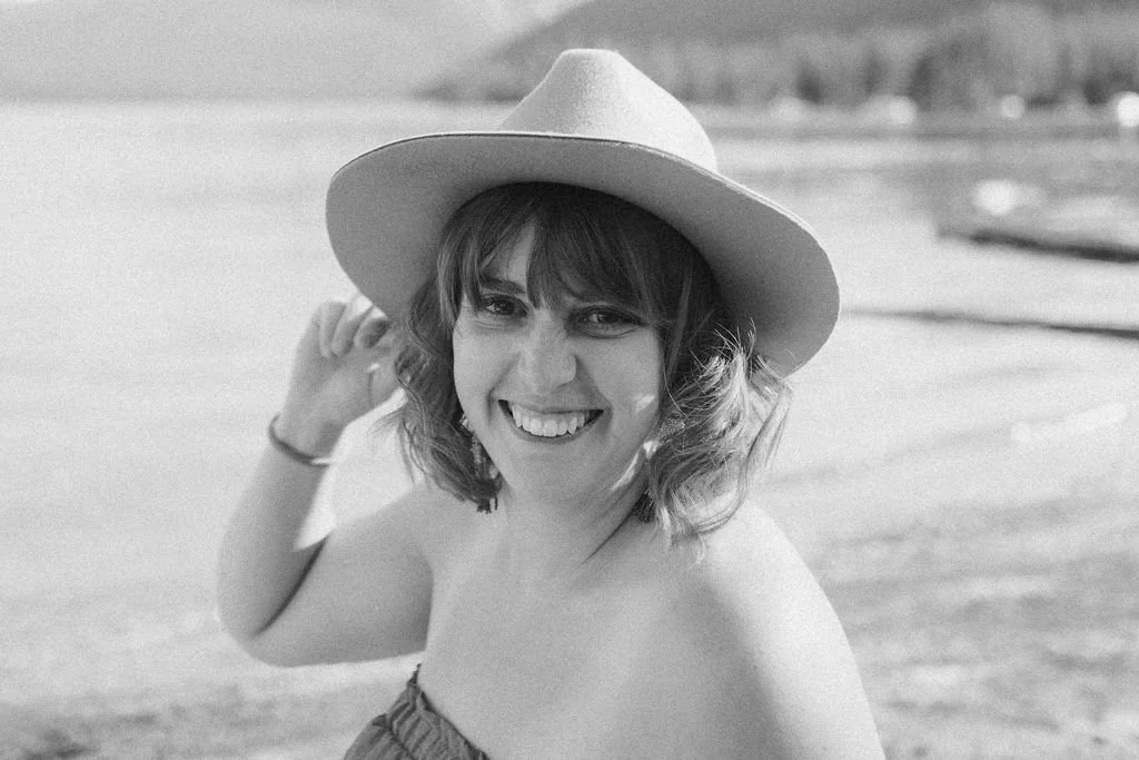 A woman smiling and wearing a wide-brimmed hat at the beach.
