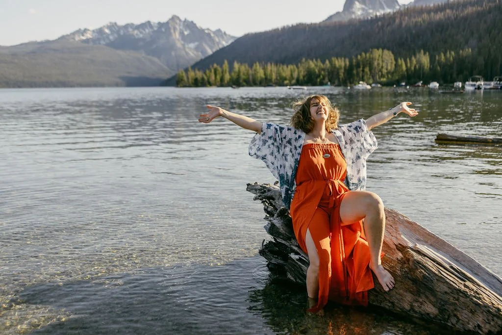 A woman in an orange dress and a patterned shawl sitting on a fallen log in a lake with her arms outstretched, smiling with her head tilted back, surrounded by mountains, trees, and a clear sky.