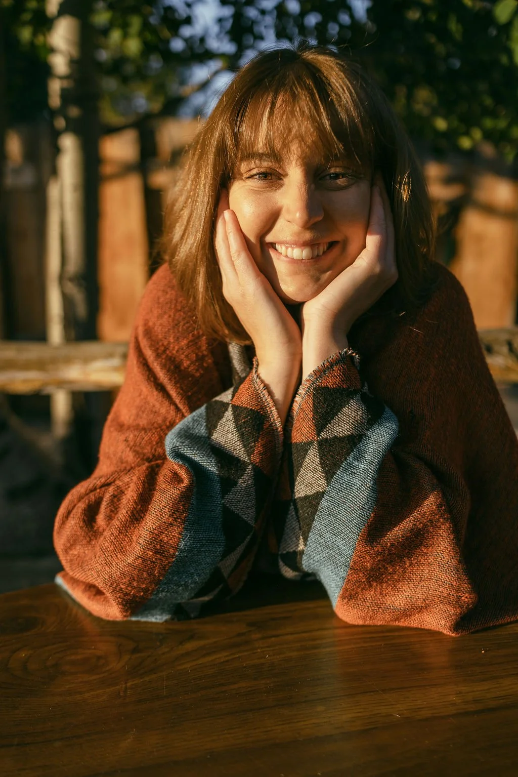 A woman with shoulder-length brown hair and a warm smile, sitting at a wooden table outdoors during sunset, wearing a rust-colored sweater and a plaid shirt.