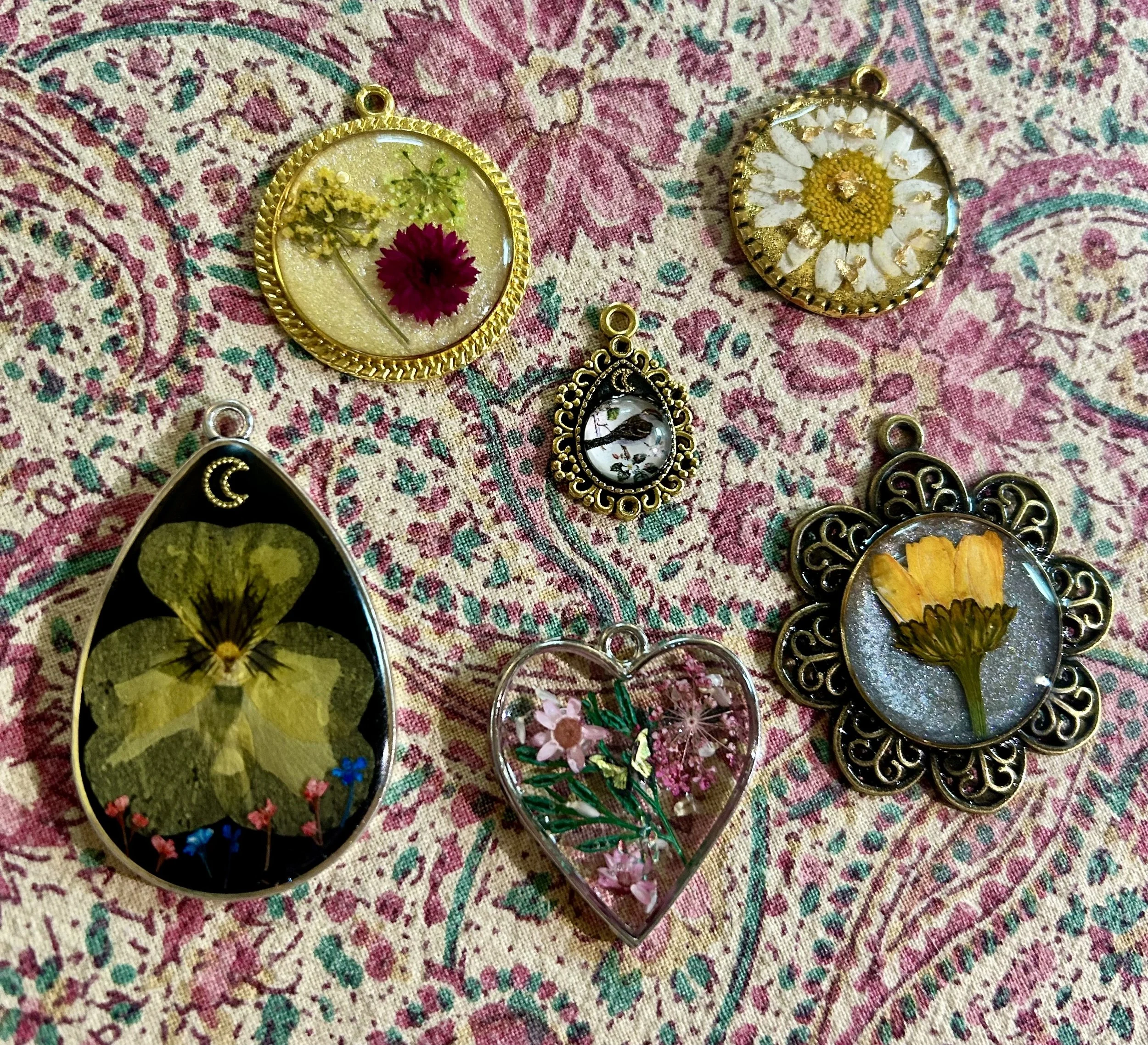 Assorted resin and metal jewelry pendants with pressed flowers and butterflies on a pink and teal patterned fabric background.