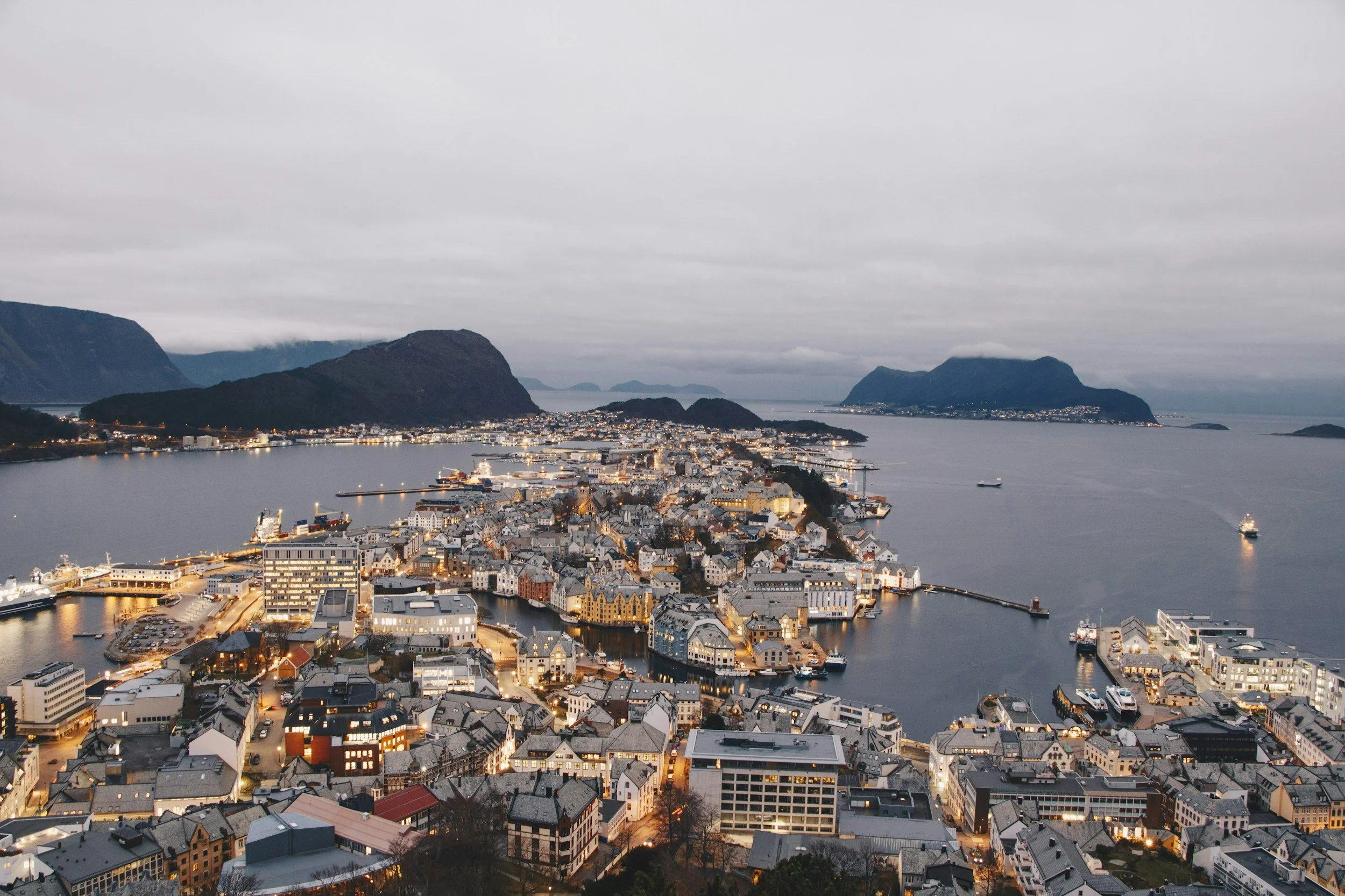 Vista de una ciudad costera en Noruega al atardecer, con edificios iluminados, montañas y aguas en el fondo