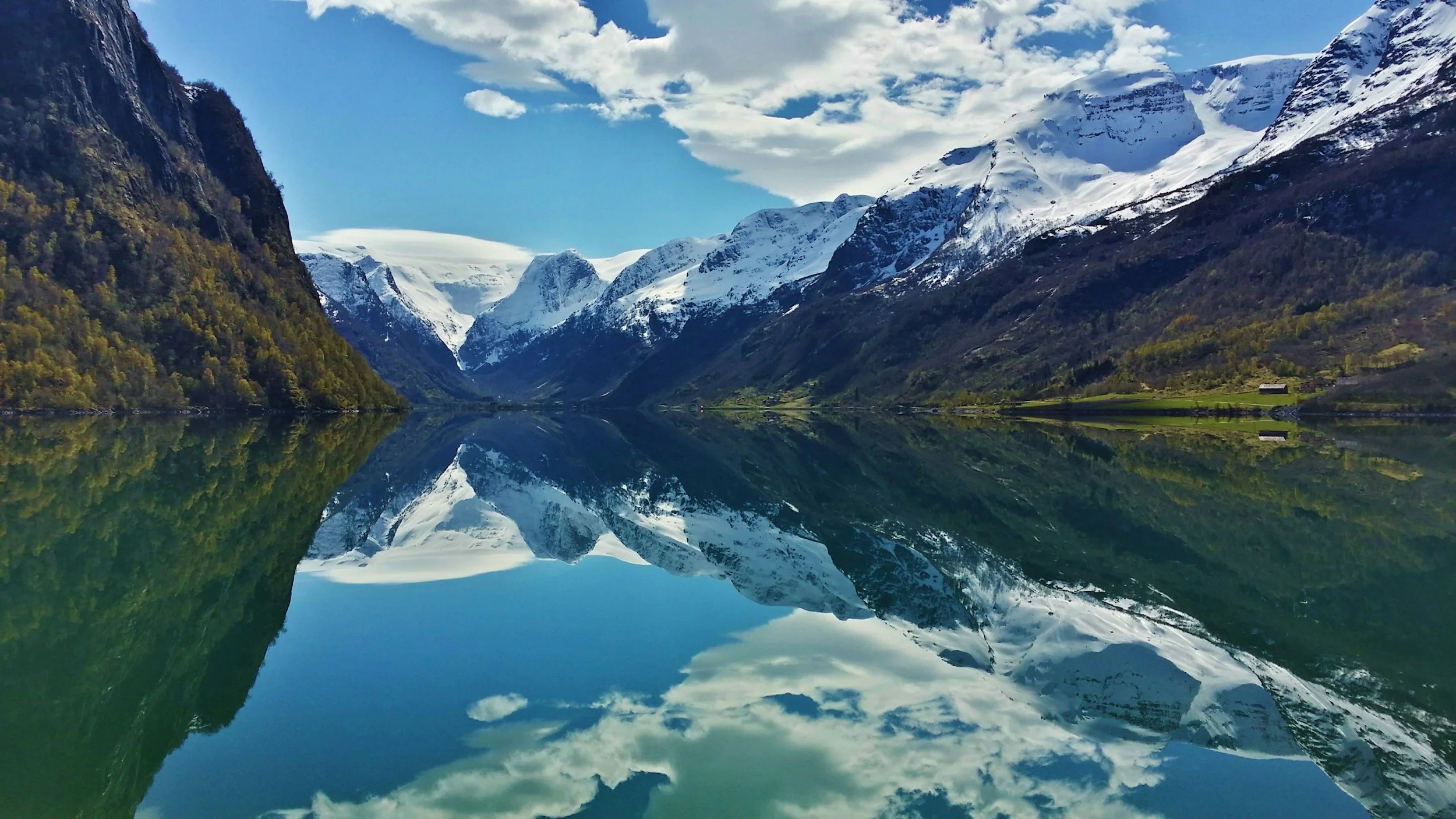 Montañas nevadas y verdes, reflejadas en un lago tranquilo con cielo azul y nubes blancas