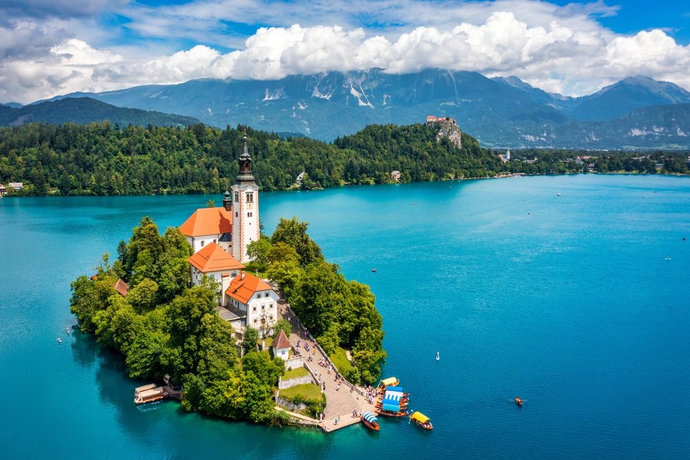 Isla con iglesia y edificios, rodeada por un lago azul, con montañas y cielo con nubes al fondo.