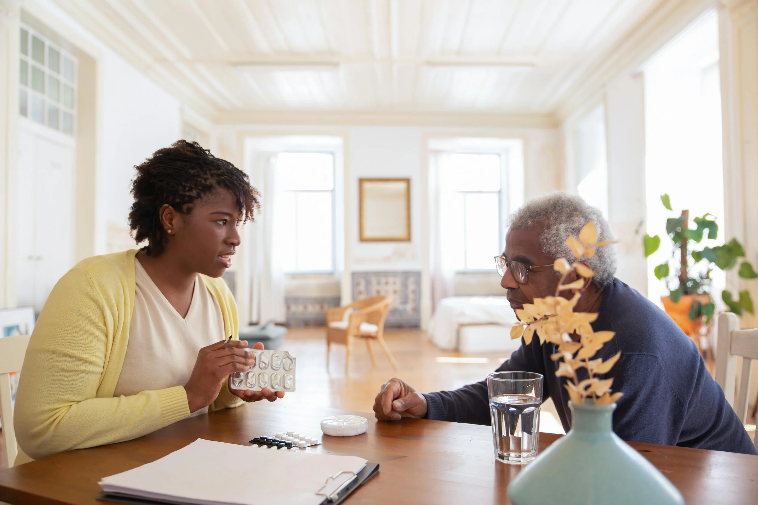 A woman and an elderly man sit at a dining table, discussing medication. The woman is holding a blister pack of pills, and there are various medication containers and a glass of water on the table. The room is bright with large windows and light-colored walls.