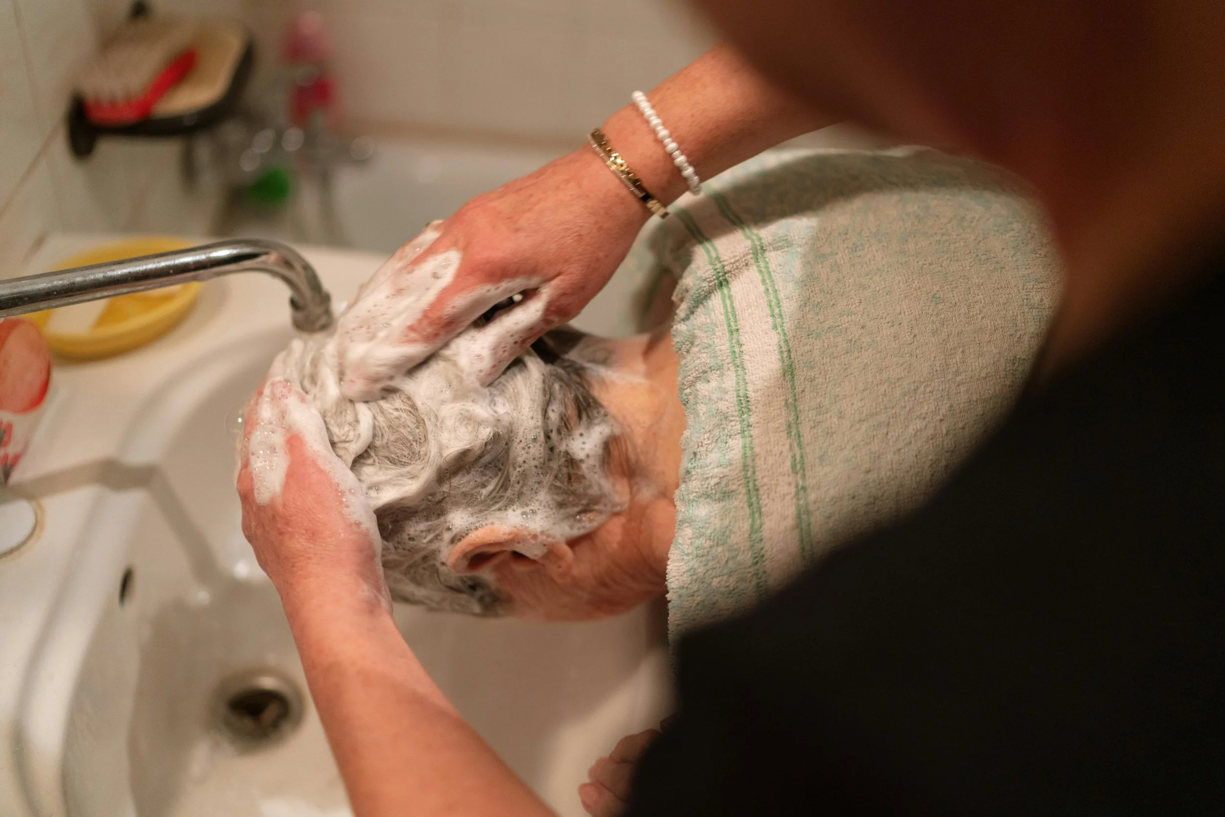Woman caregiver washing another person's hair over a kitchen sink, using soap and creating foam, with a towel draped over the person's shoulders.