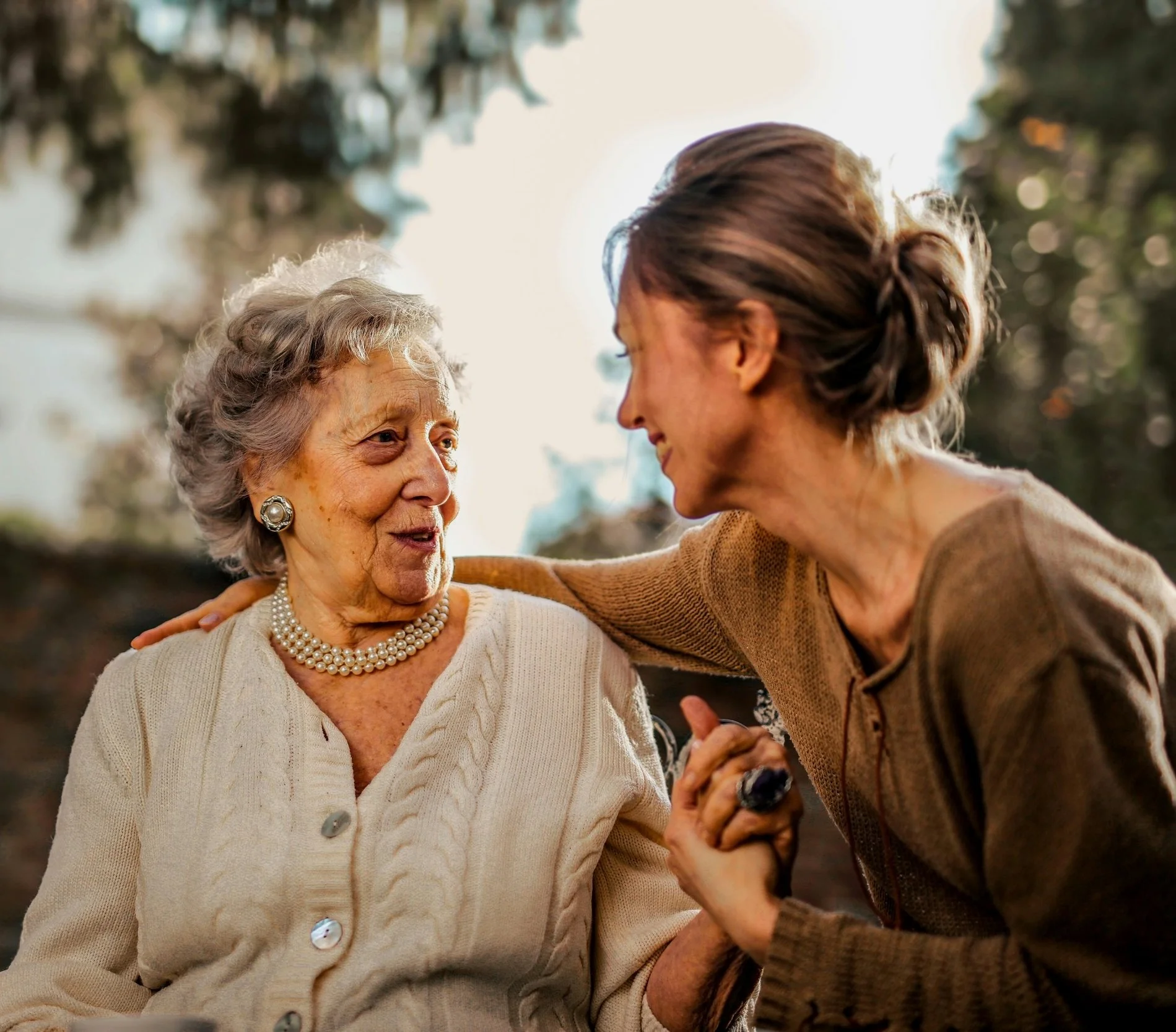 An elderly woman and a middle-aged woman caregiver are smiling and holding hands, having a heartfelt conversation outdoors during daylight.