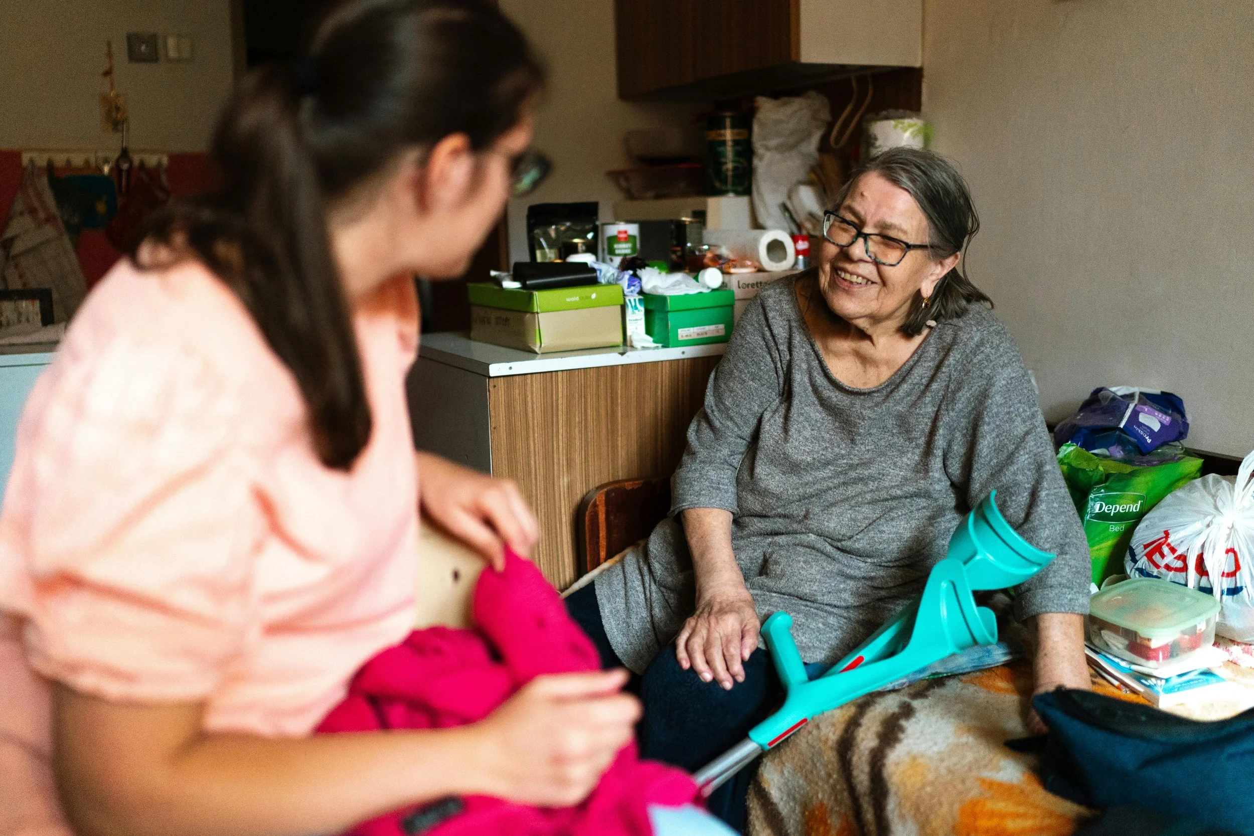 An elderly woman sitting on a table, smiling at a caregiver who is holding a sweater, in a cluttered room with various household items around.