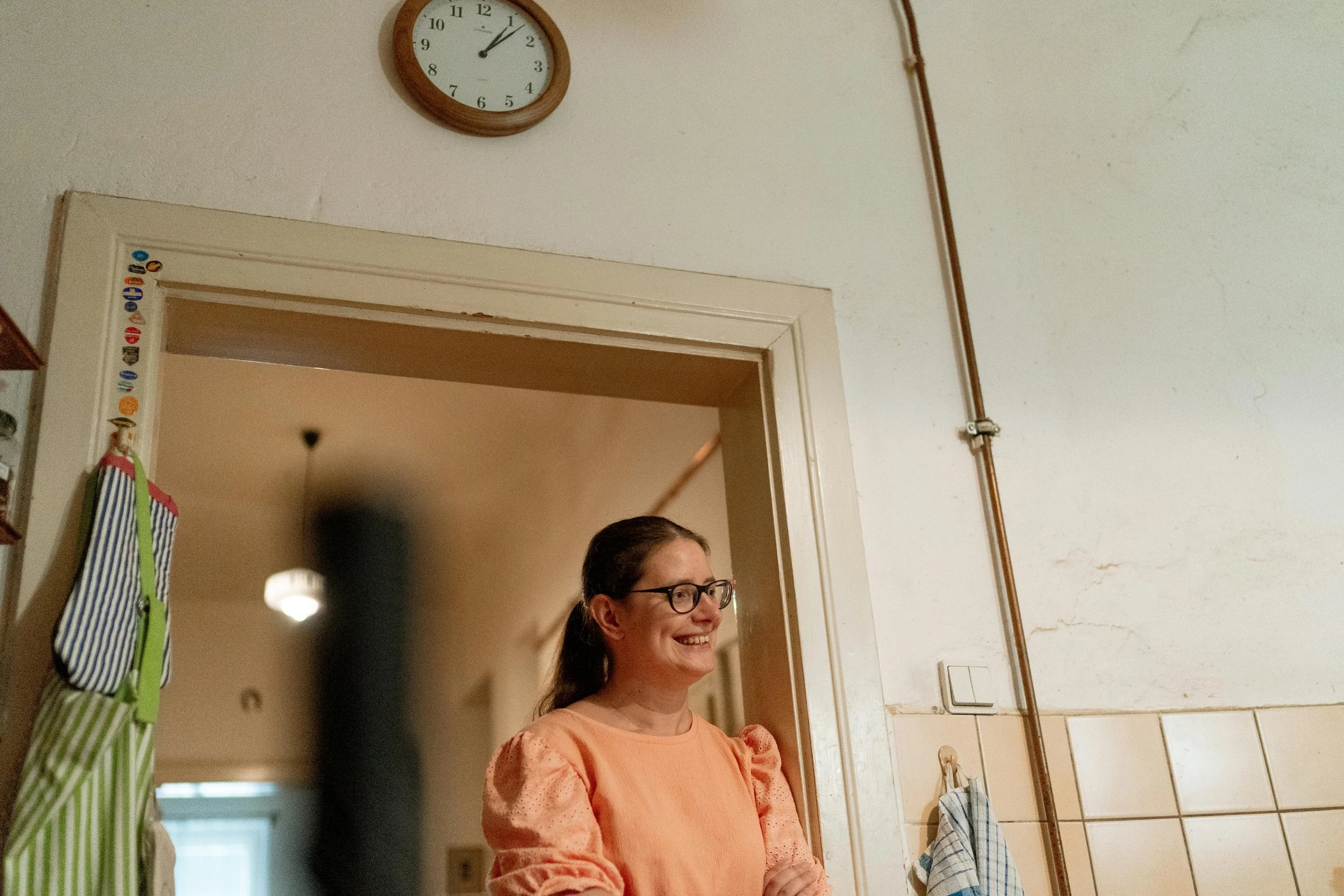 A woman caregiver with glasses and long brown hair smiling, standing in a doorway in a kitchen. The wall above has a round clock.