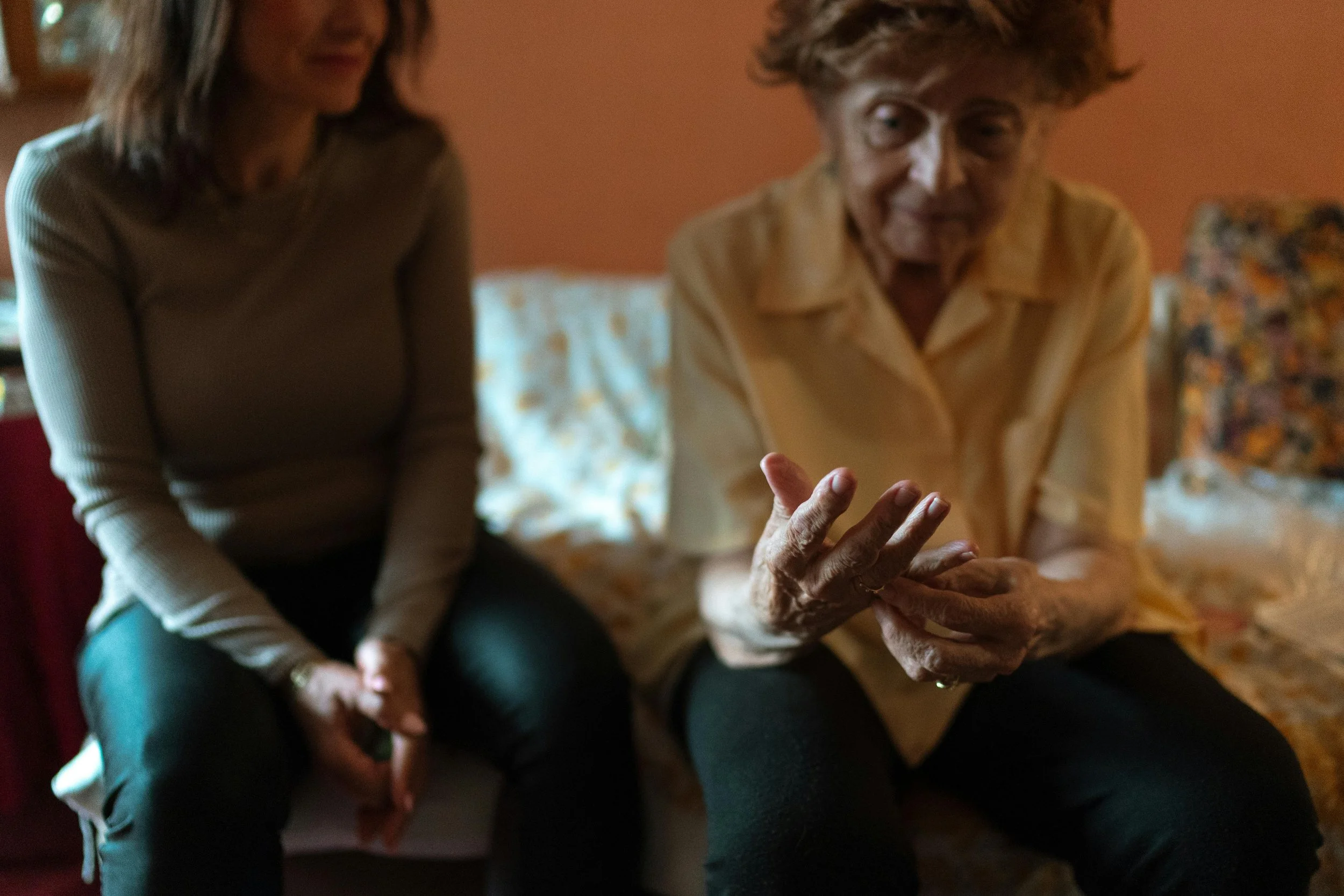 An woman caregiver and a younger woman sitting on a bed in a dimly lit room, engaged in conversation, with the elderly woman gesturing with her hands.