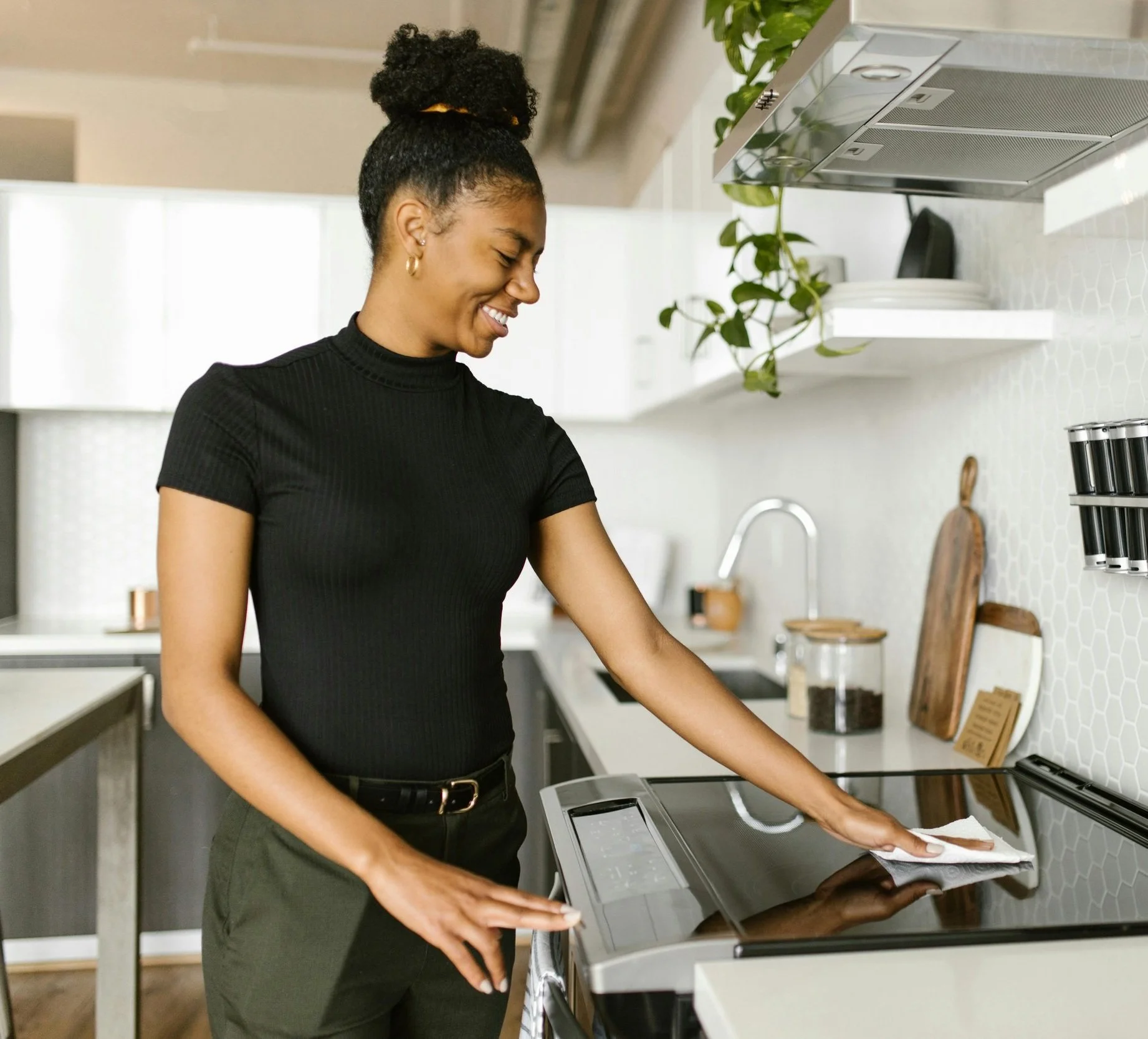 A woman caregiver in a black turtleneck and black pants cleaning an electric stove in a modern kitchen with a white hexagonal tile backsplash.