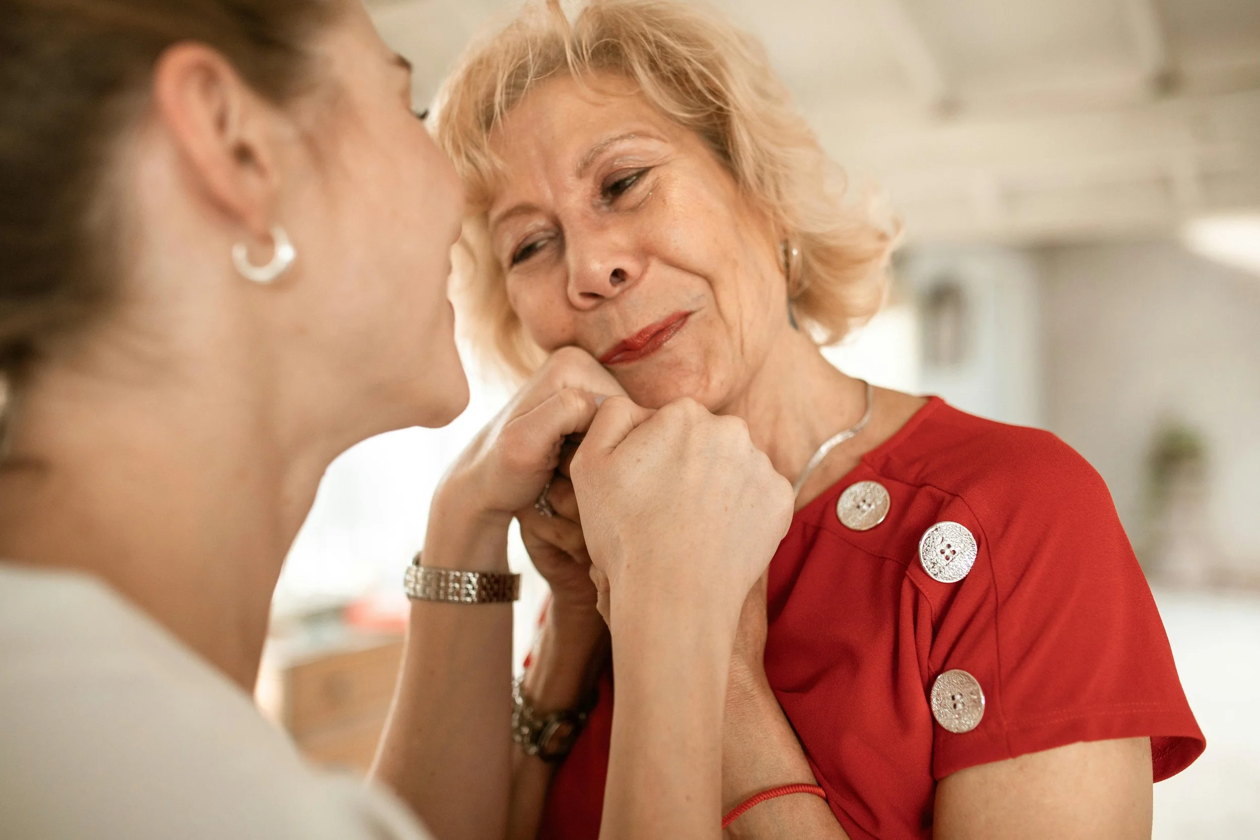 An elderly woman with blonde hair wearing a red top with silver buttons is holding hands with a caregiver, touching foreheads in an emotional moment.