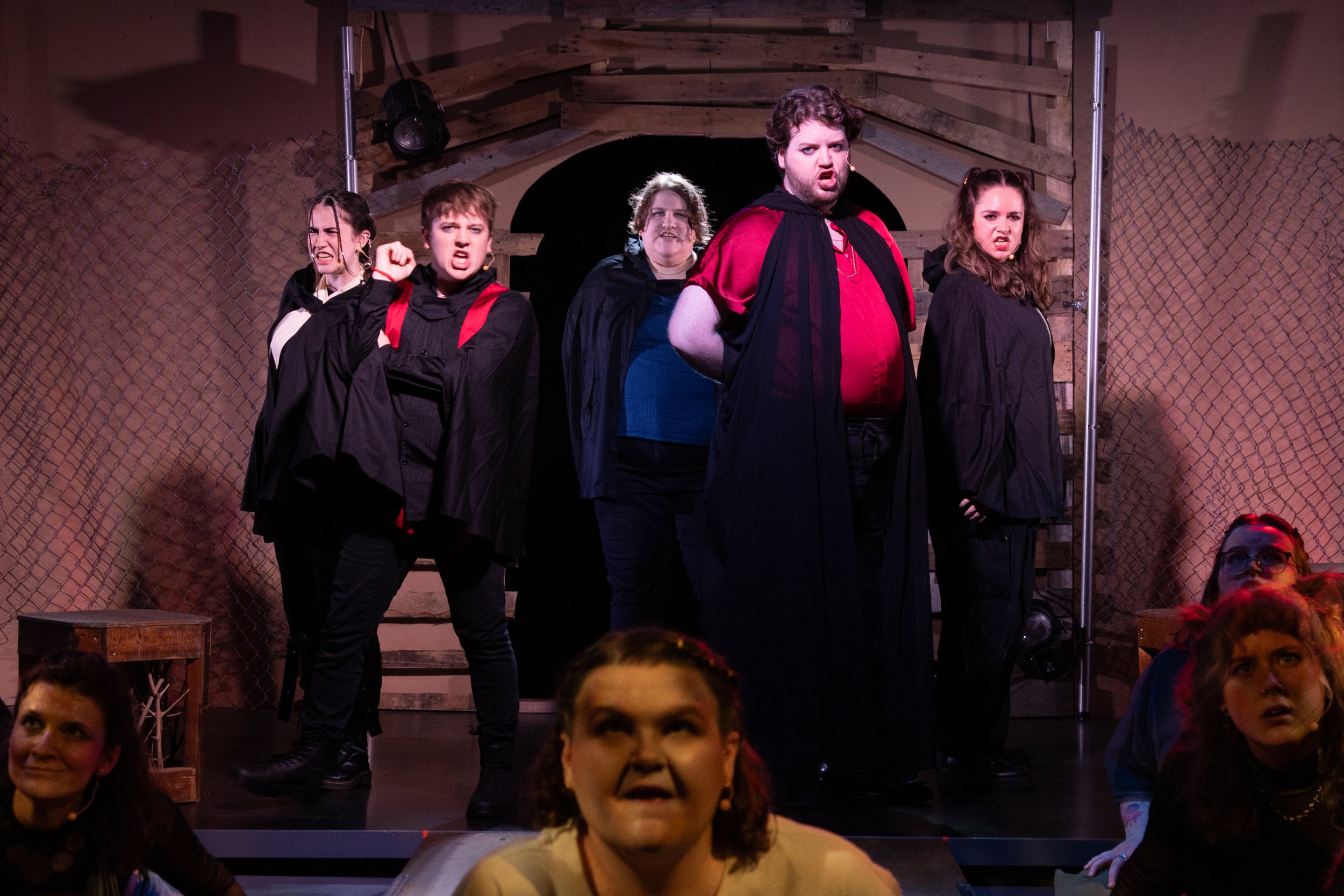 Actors on stage performing a play with serious and intense expressions, dressed in dark clothing and cloaks, set against a backdrop resembling a wooden fence and archway.
