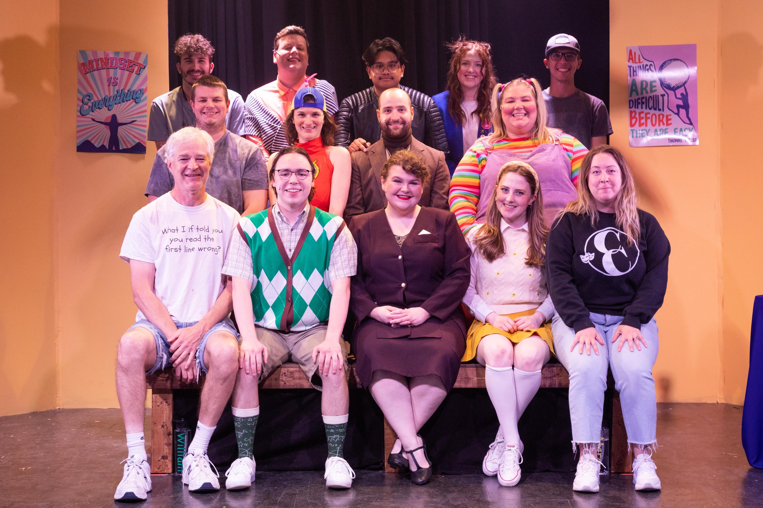 Group of actors on stage in a school play, with three rows of performers and colorful posters on yellow and black walls.