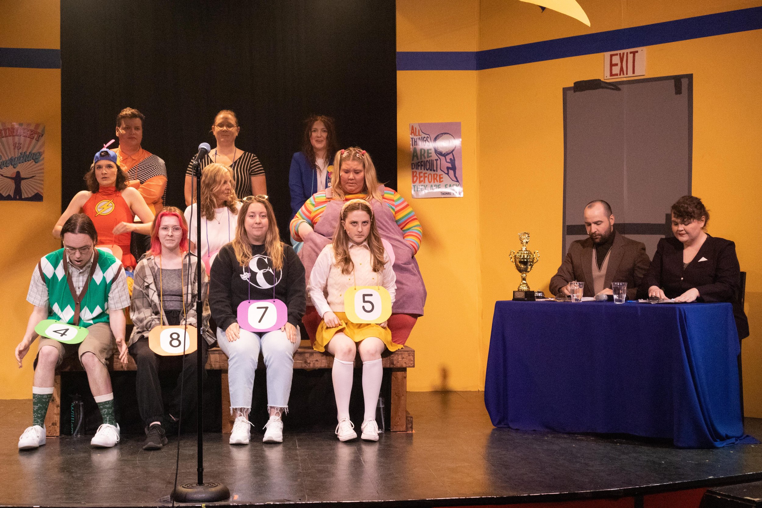 A group of young people with colorful costumes and numbered signs sitting on a stage, with some standing behind them, and two adults sitting at a table to the right, with a trophy and glasses of water, in a yellow-walled room with posters.