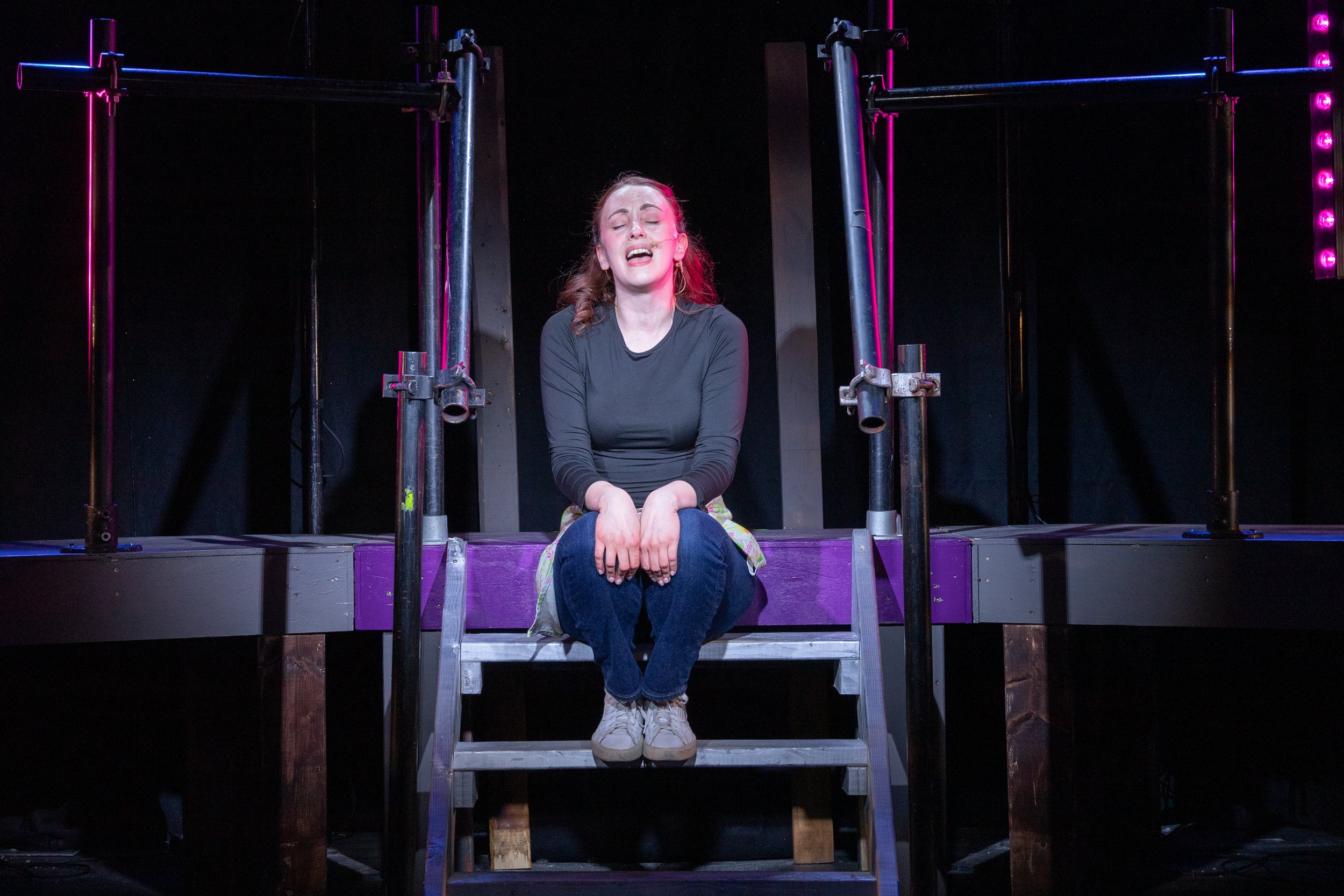 A young woman in black shirt and jeans sitting on a small gray platform with a distressed expression, on a stage with black background and purple neon lighting.