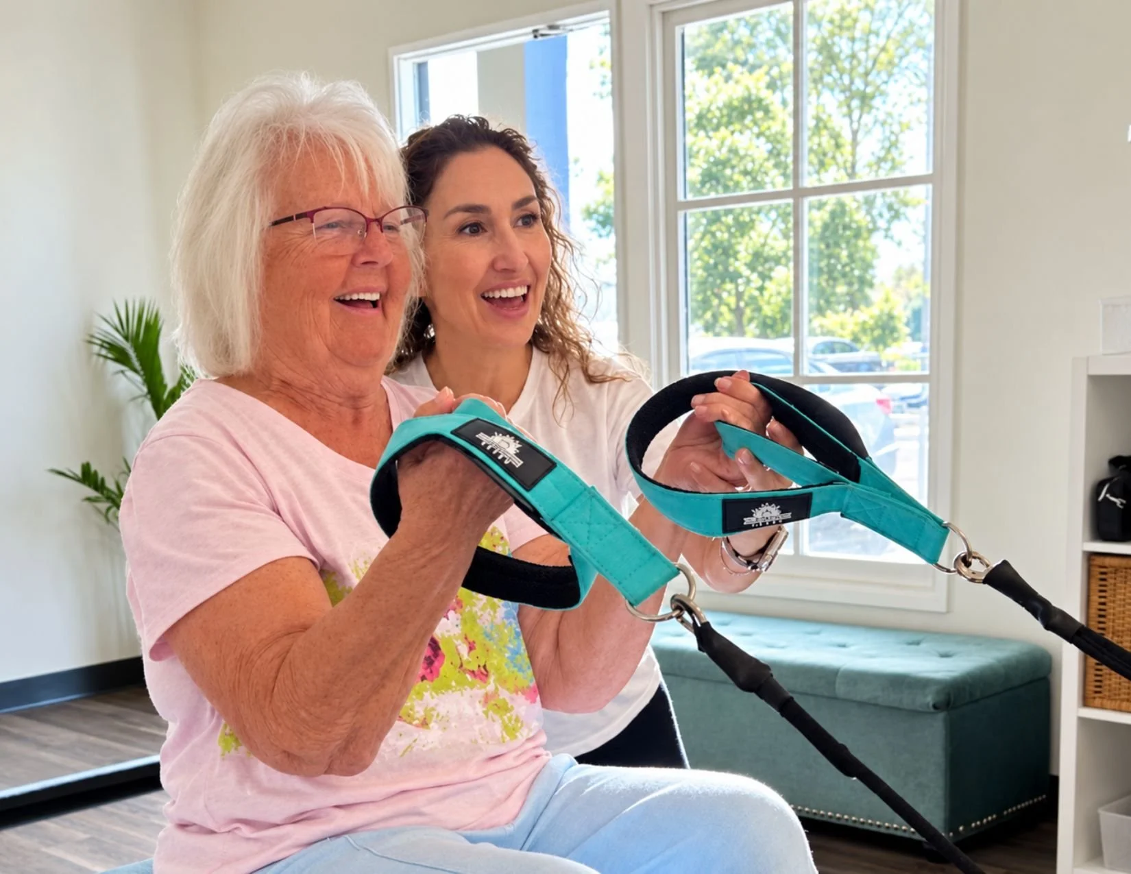 A smiling woman with white hair sits on a Pilates Reformer holding the straps while a female specialist leans in beside her, guiding the movement.