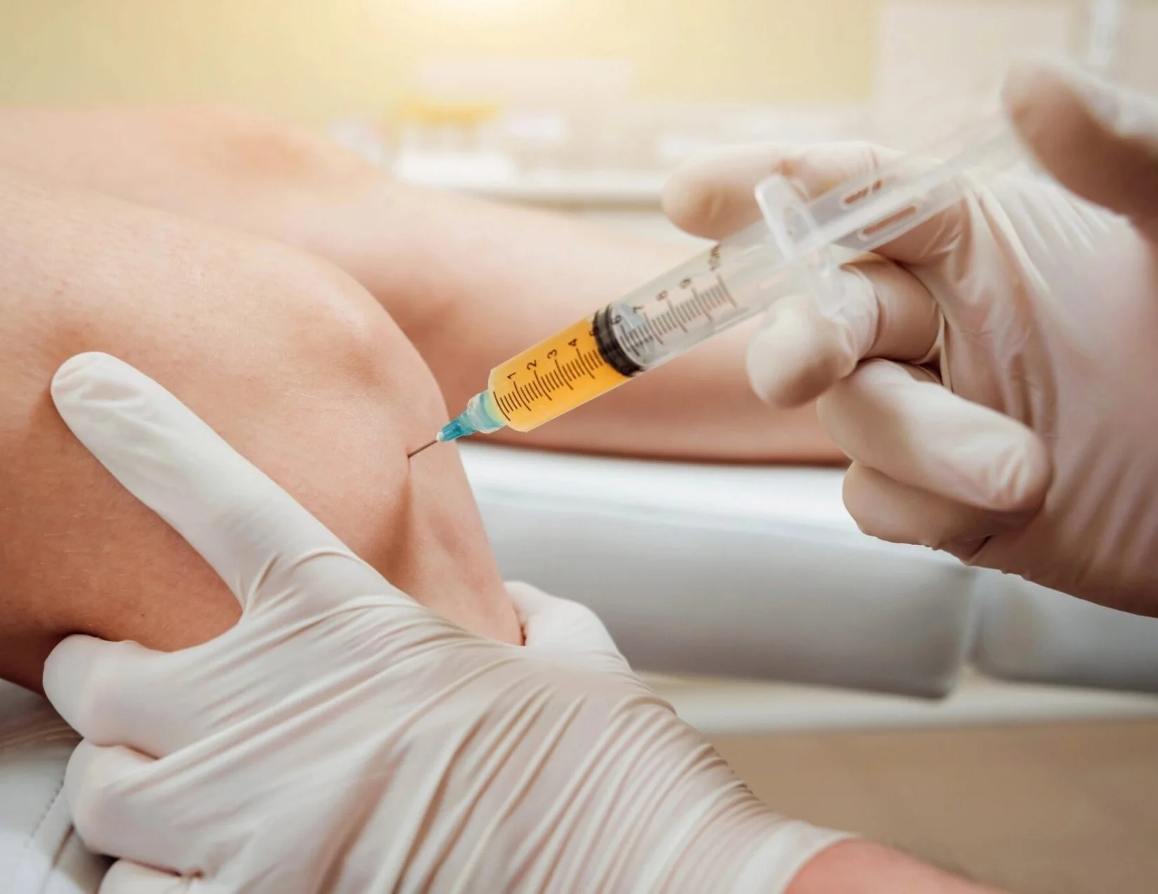 Gloved hands inserting a needle into the side of a knee, the syringe filled with a clear yellow fluid.