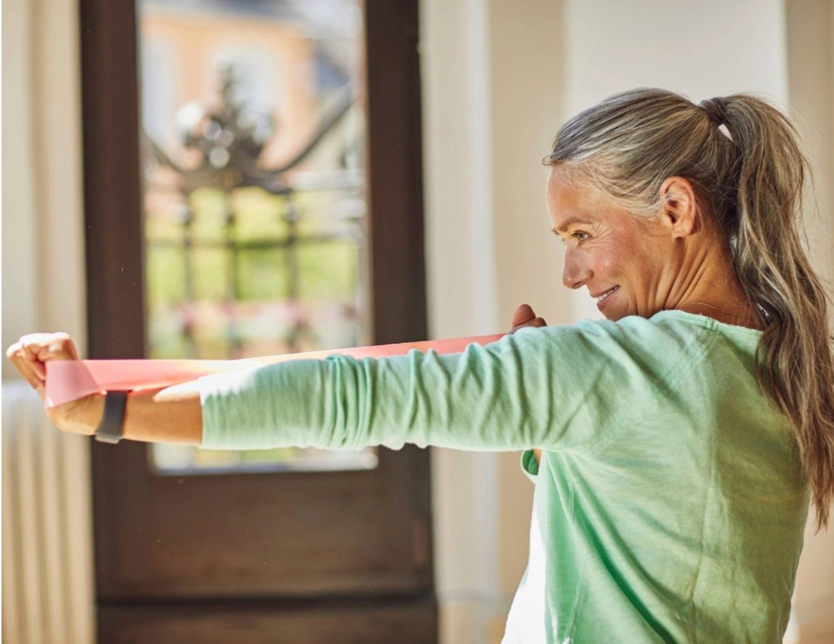 A woman with a grey ponytail smiling as she pulls a pink resistance band back with one hand, her other arm stretched straight out in front of her.