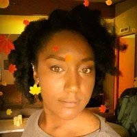 A young woman with natural curly hair, wearing a gray top, yellow star-shaped earrings, and a small red star sticker on her forehead, standing in an indoor setting with warm lighting.