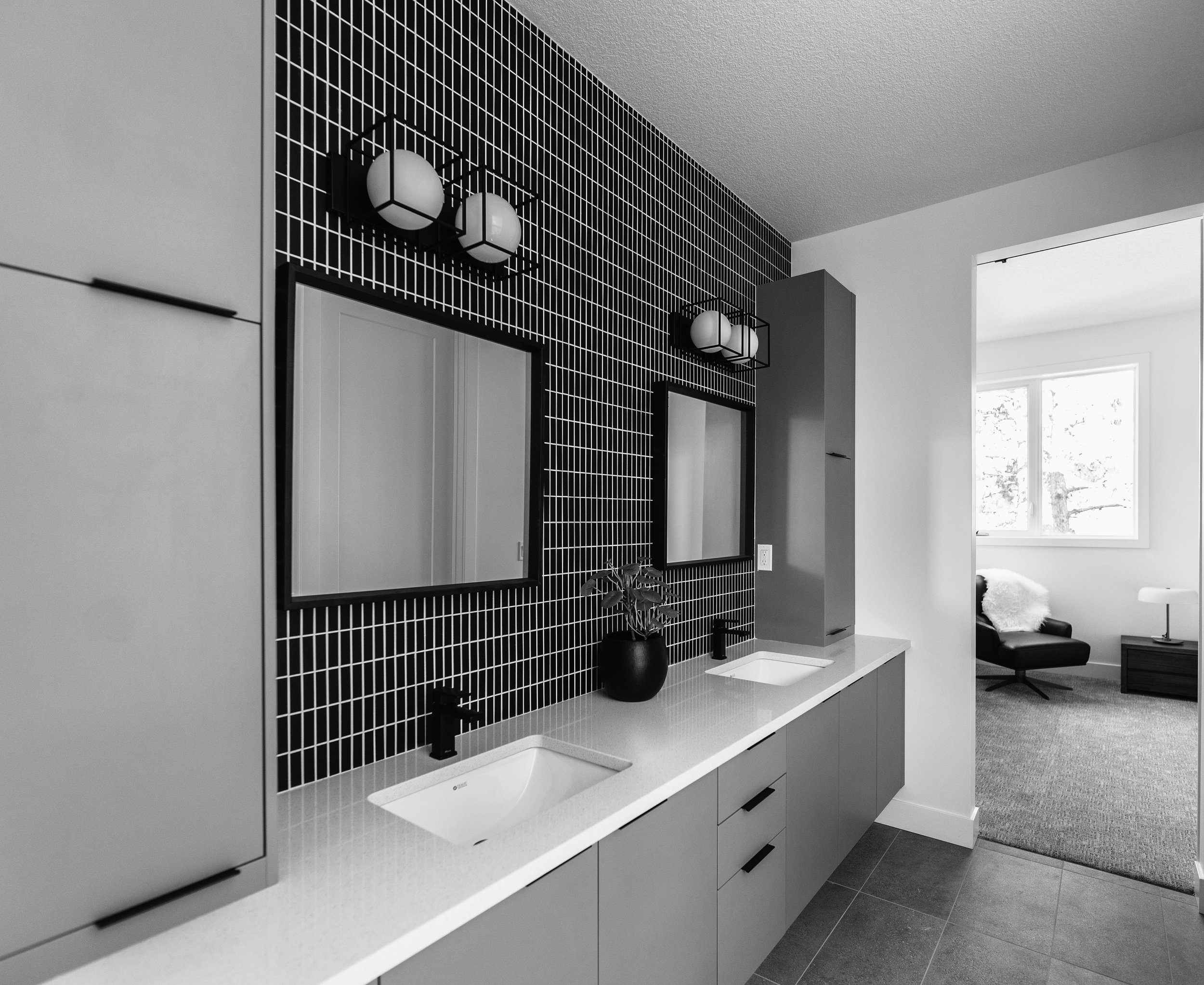 Modern bathroom with black tiled accent wall, two rectangular mirrors, two black sinks with black faucets, and a white countertop. There are two wall-mounted light fixtures above each mirror, and a small black vase with a plant on the counter. An open doorway reveals a living room with large windows, a black chair, and a side table with a lamp.