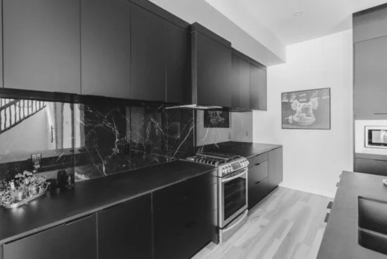 Modern black kitchen with black cabinets, black marble backsplash, stainless steel oven, and wooden flooring.