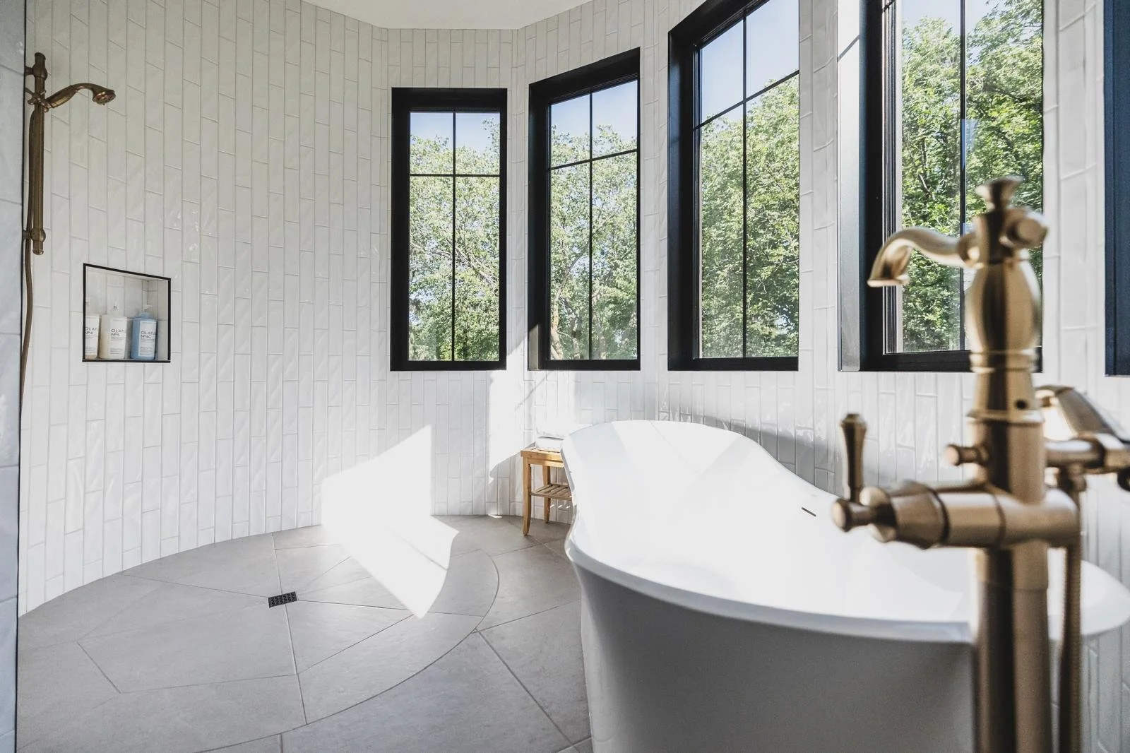 Modern bathroom with white tiled walls, black-framed windows letting in natural light, a white freestanding bathtub, wooden stool, and brass fixtures.